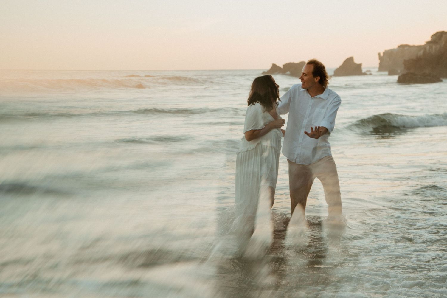 Couple in white outfits standing in ocean waves at sunset on a scenic beach.