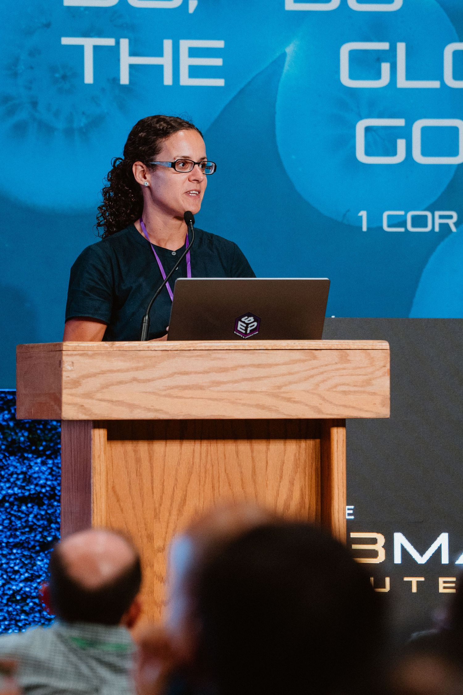 Woman presenting at a podium with a laptop at a conference with a blue biblical scripture backdrop.