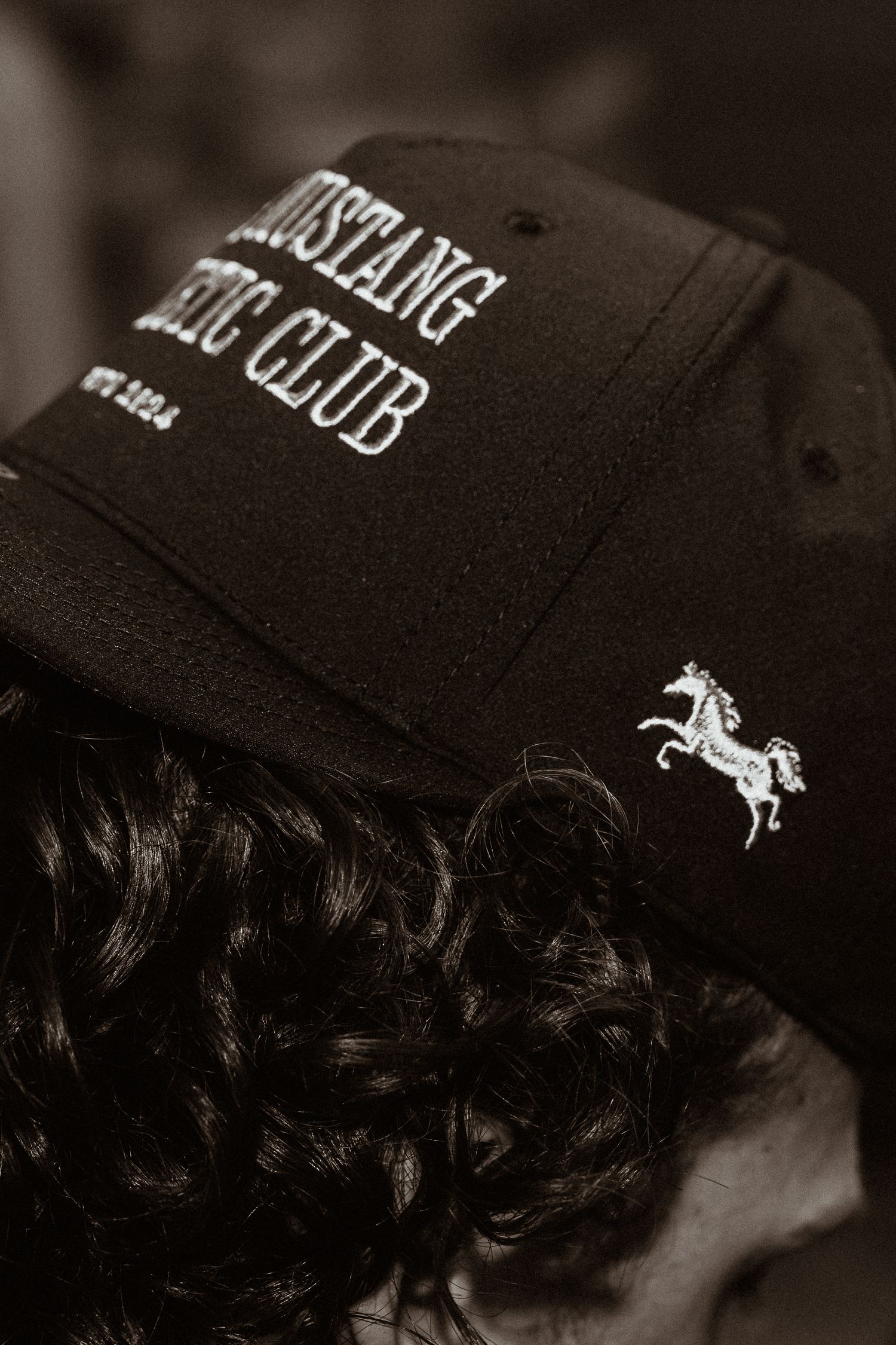 Black Mustang Drug Club embroidered baseball cap with horse logo on curly hair.