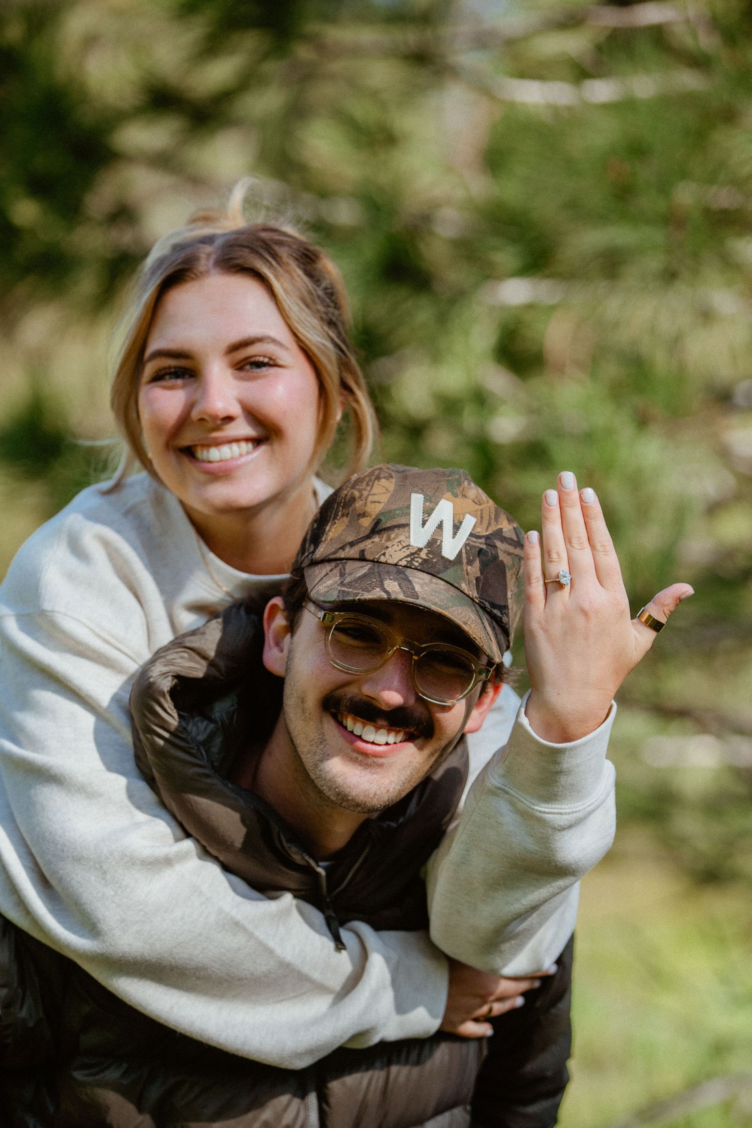 Joyful newly engaged couple, woman showing ring while getting a piggyback ride outdoors.