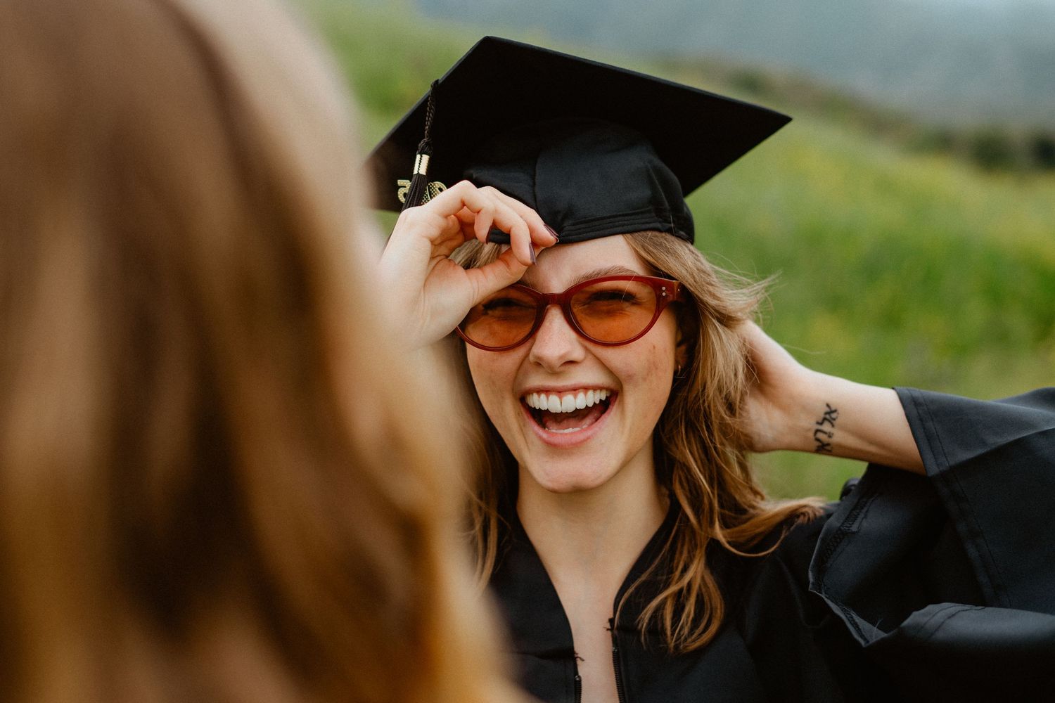 Laughing graduate in black cap holds orange sunglasses while celebrating in a green meadow.