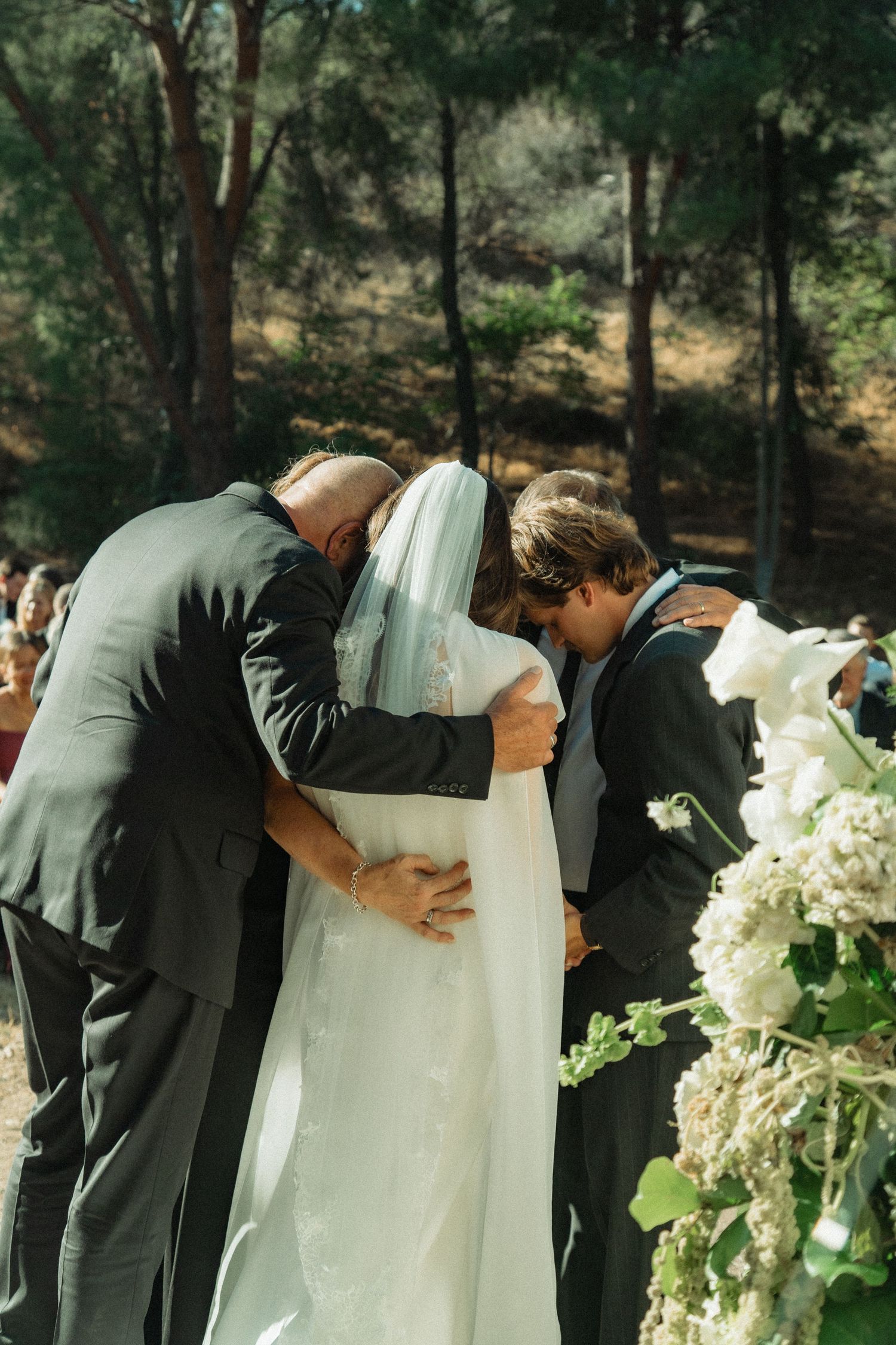 Three people sharing emotional group hug at outdoor wedding altar surrounded by white floral arrangements.