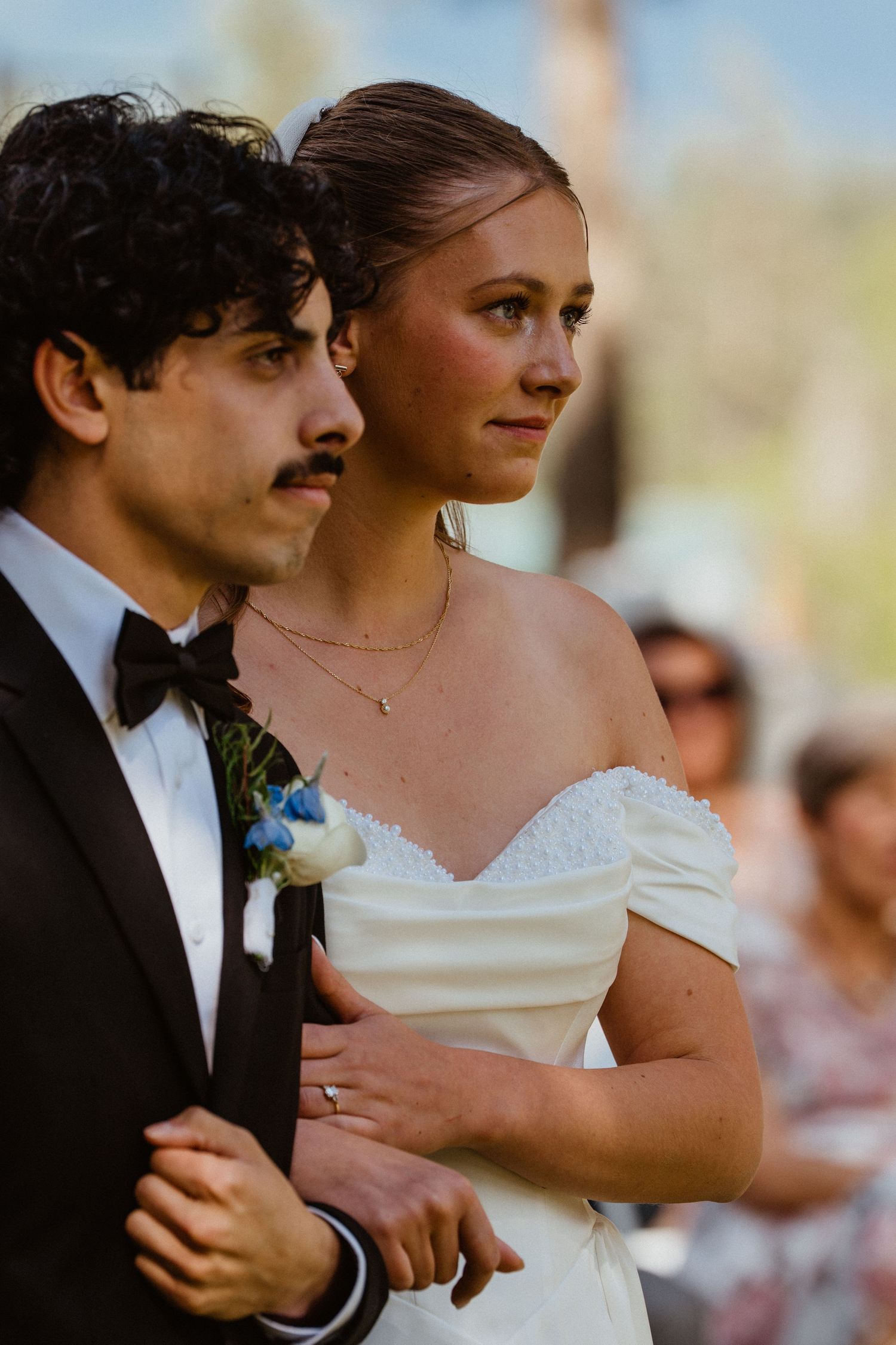 Bride in off-shoulder gown and groom in black tuxedo standing together during outdoor wedding ceremony.