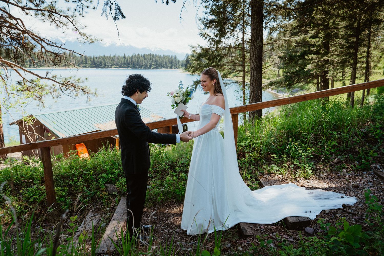 Couple exchanges vows outdoors by a mountain lake, bride in white gown, groom in black suit, surrounded by pine trees.