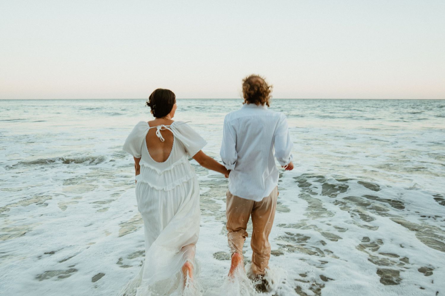 Couple in white wedding attire holding hands walking along ocean shore waves at sunset beach.