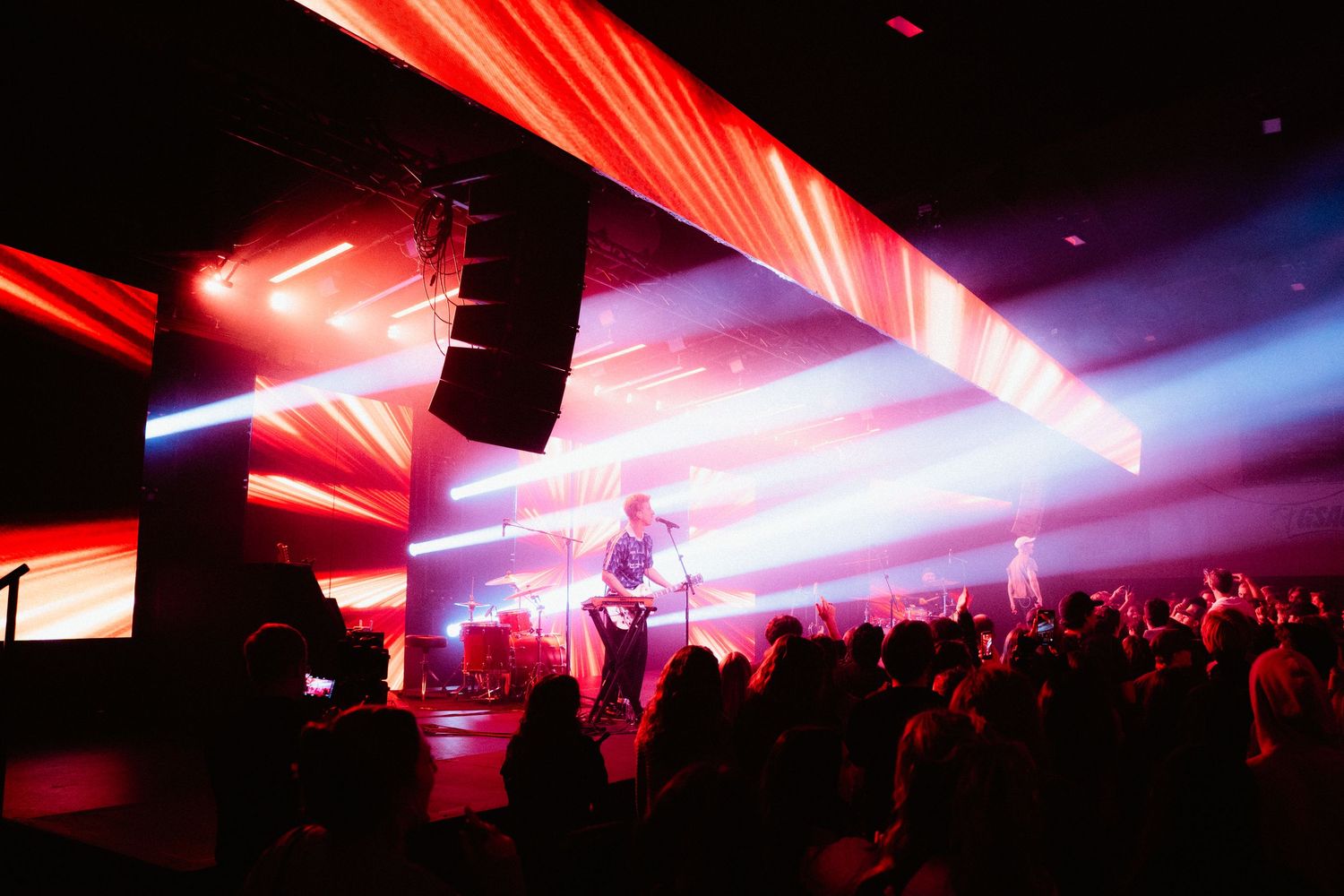 Band performing on stage with dramatic red and white light beams over an enthusiastic crowd.