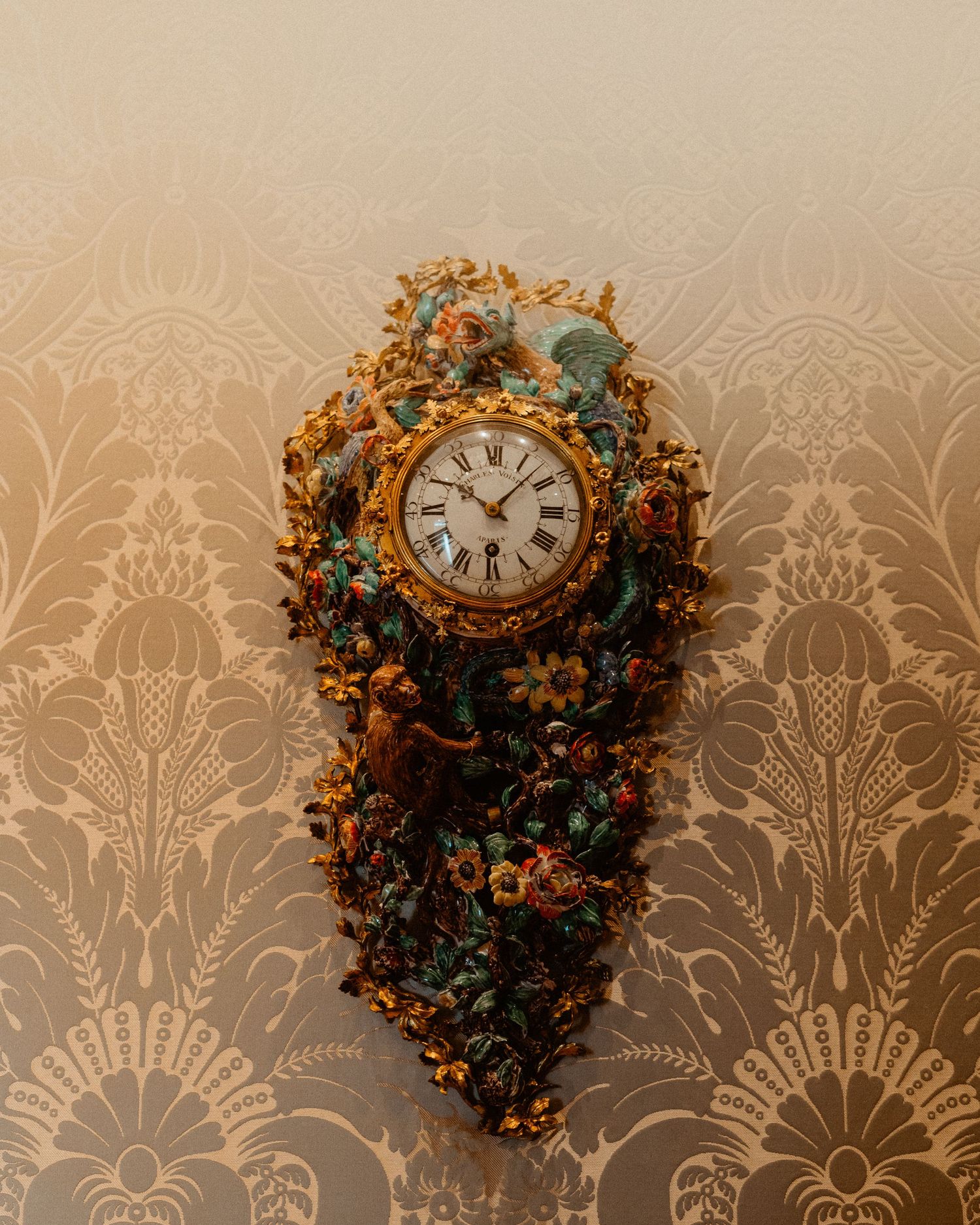 Ornate antique wall clock decorated with colorful floral motifs in gold against damask patterned wallpaper.