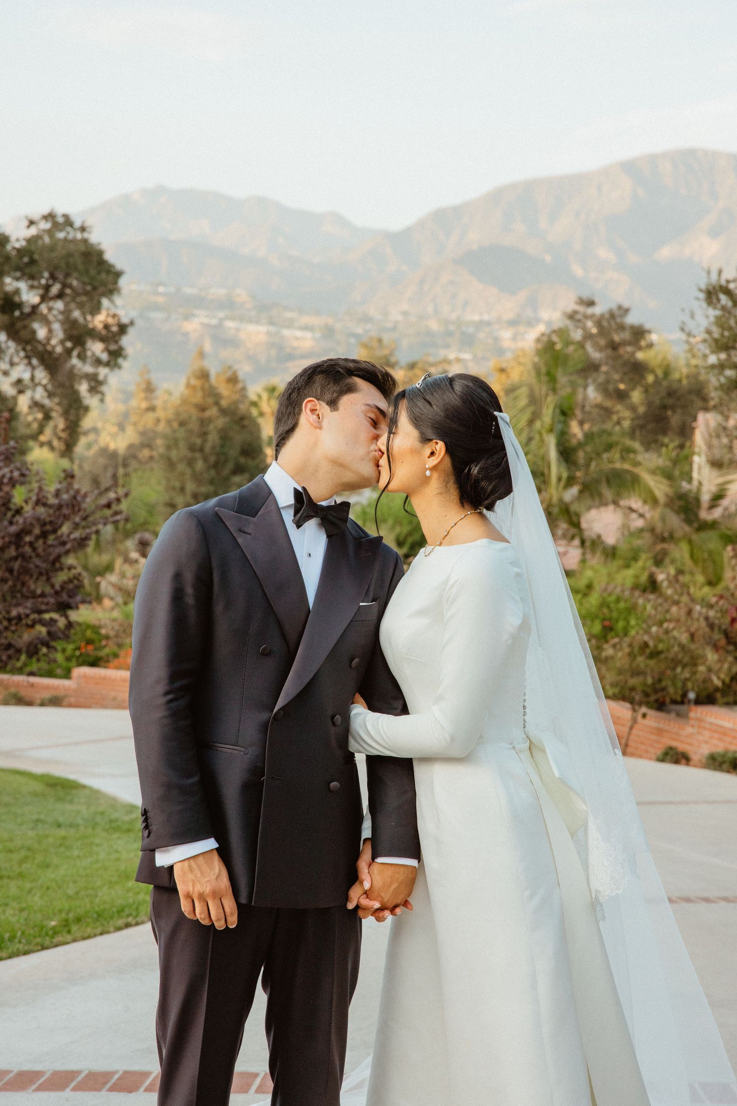 Bride and groom share a kiss while holding hands with scenic mountain landscape behind them outdoors.