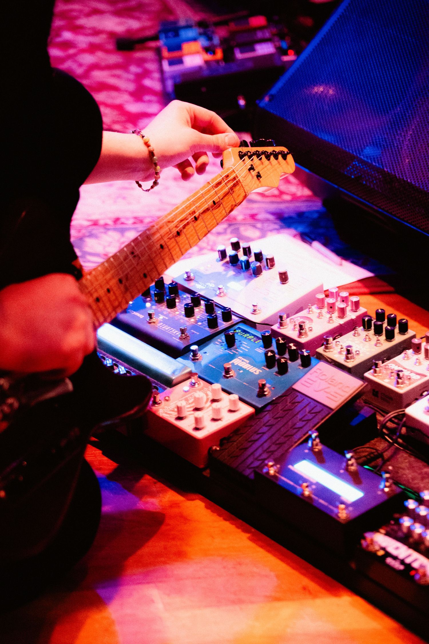 Guitarist adjusting guitar near effects pedal board on stage during live concert performance.