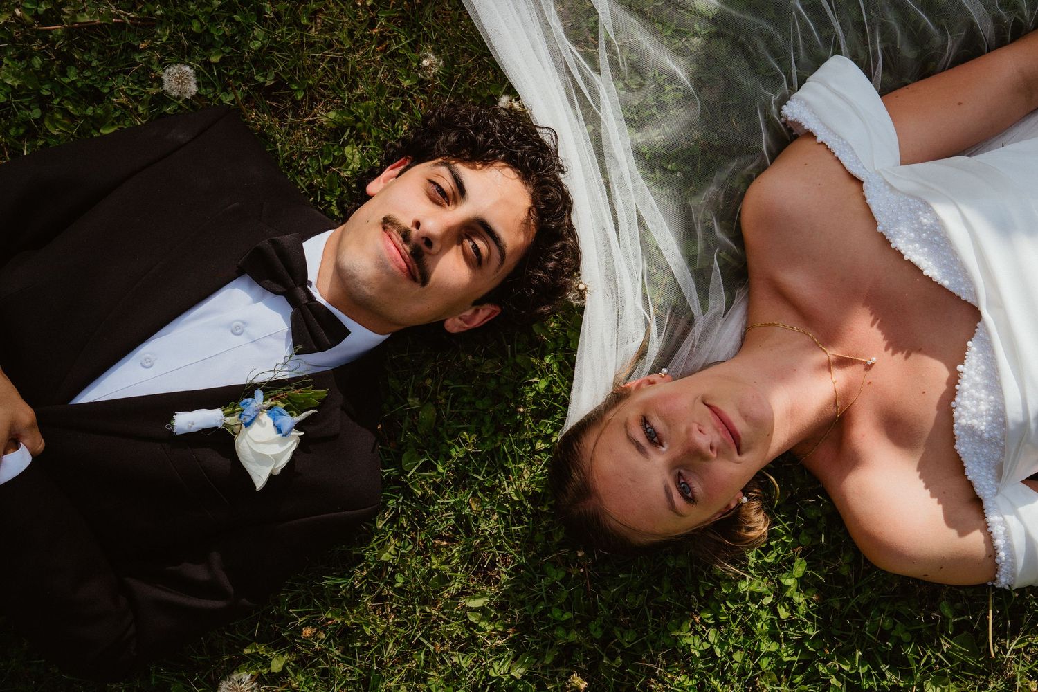 Bride and groom lying on green grass looking up, wearing wedding attire with veil spread around them.