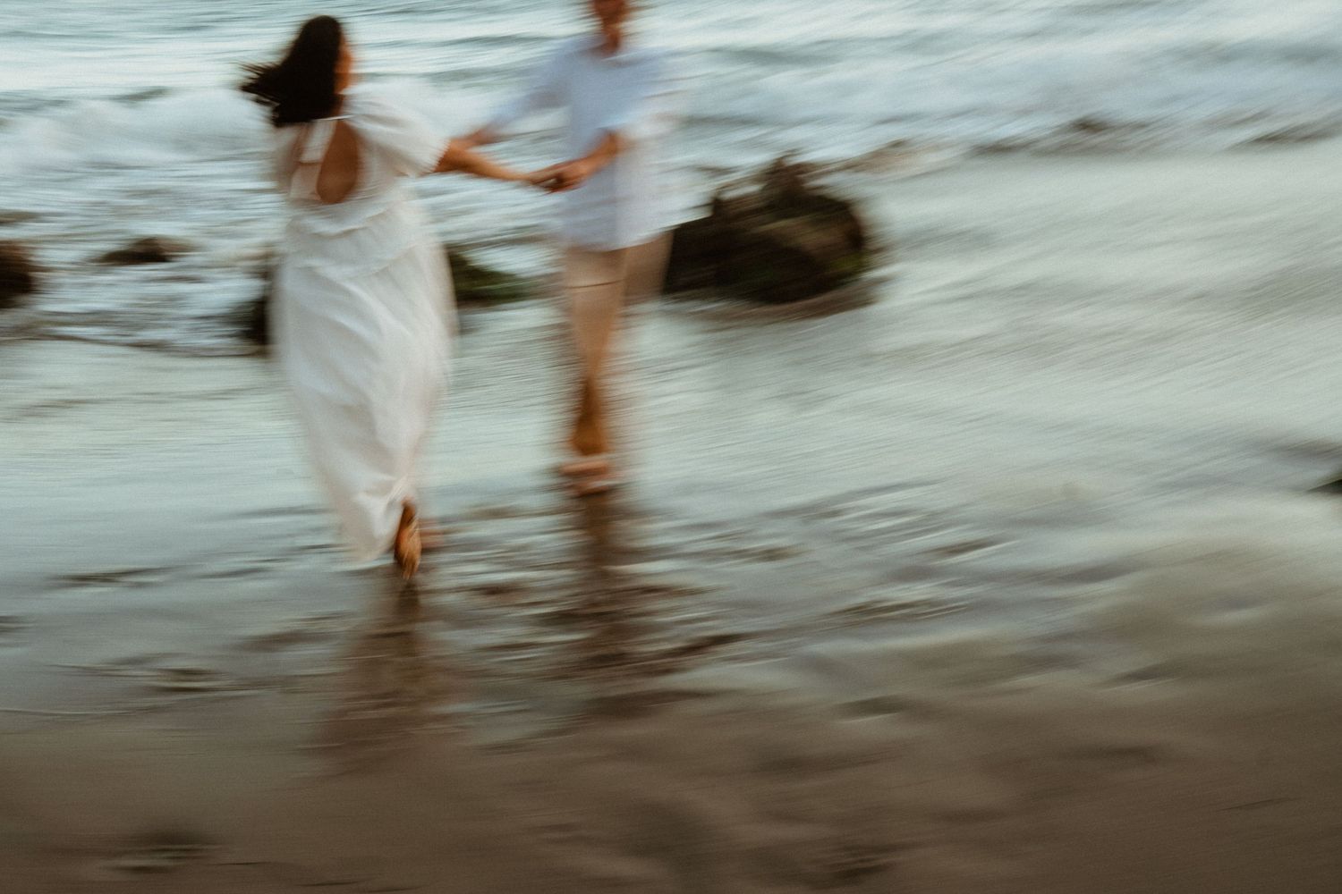 Couple in white attire running barefoot on a sandy beach shore with ocean waves, captured with motion blur effect.