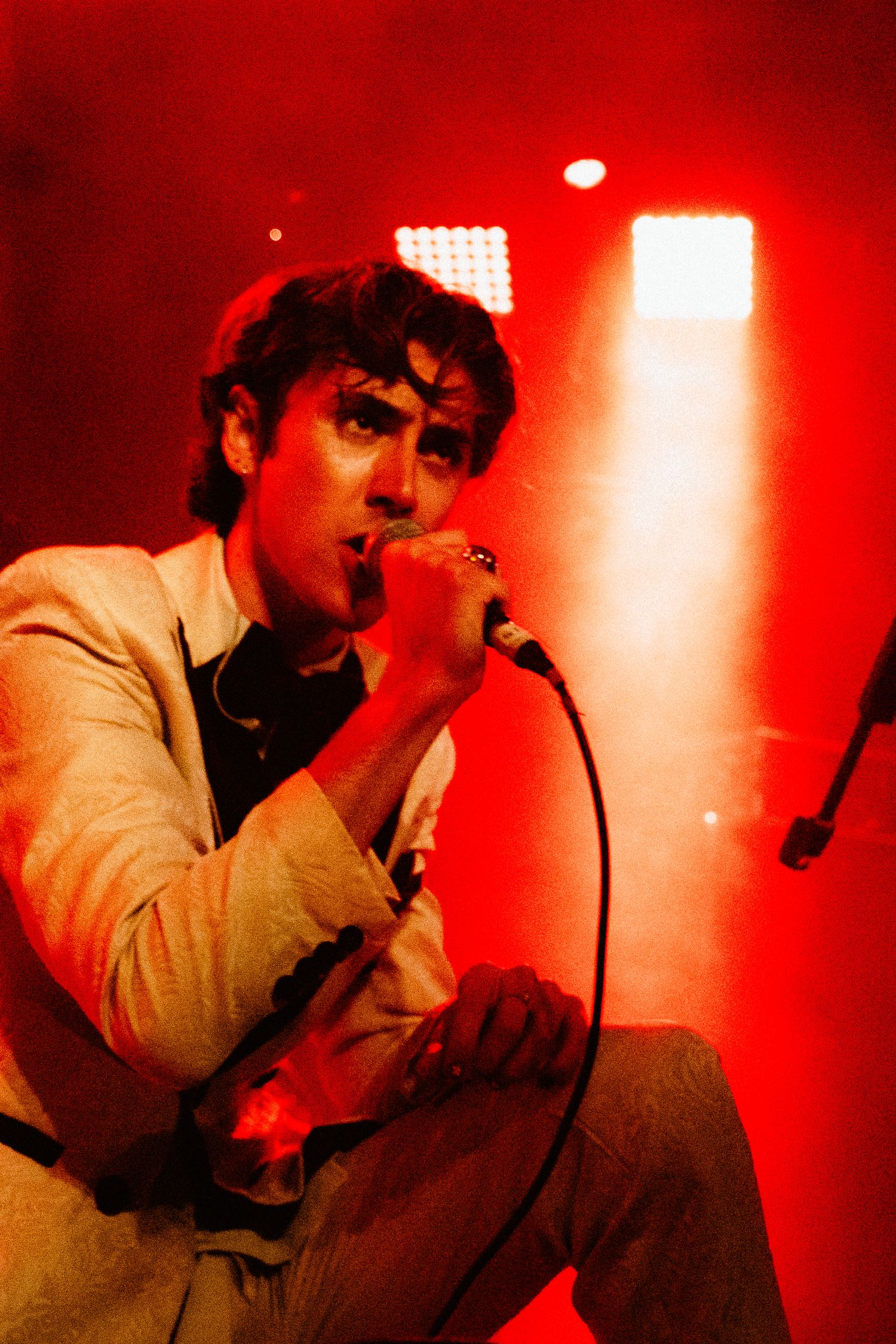 Male vocalist in white blazer singing passionately into microphone under red stage lighting.