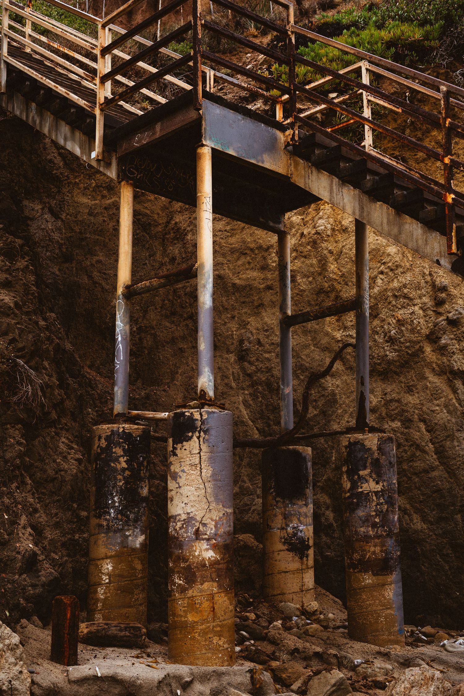Weathered wooden staircase supported by rusty metal pillars embedded in a rocky coastal cliff face.