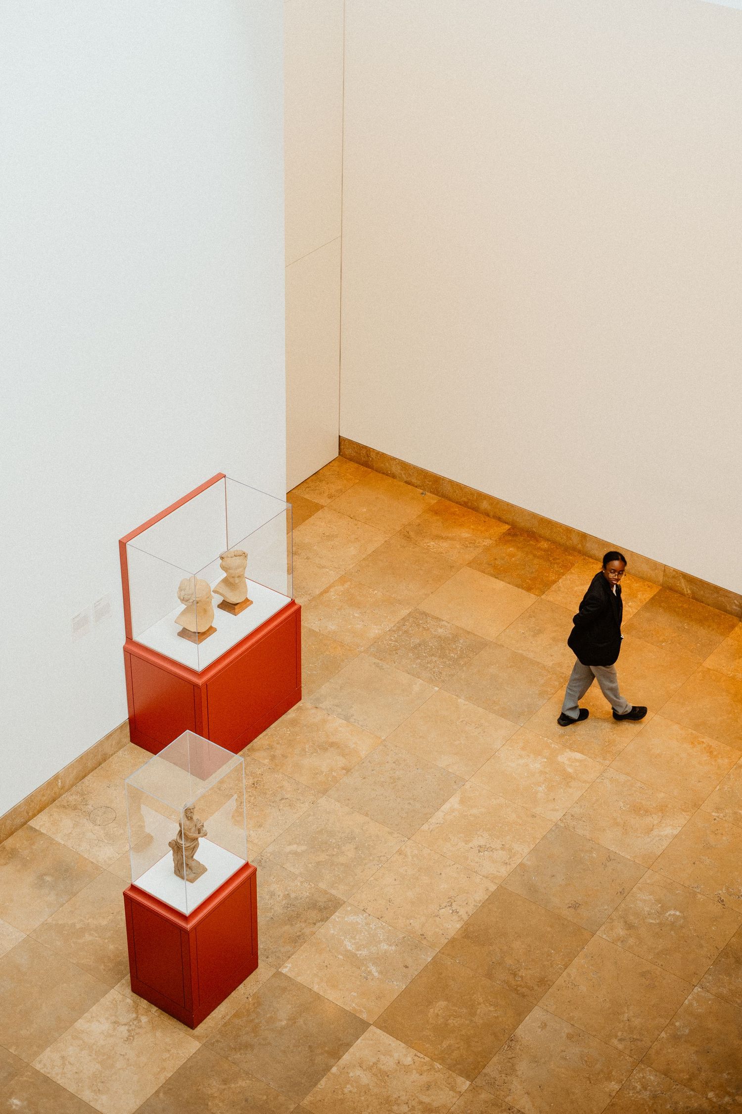 Aerial view of museum gallery visitor walking past ancient sculptures displayed in red pedestals on travertine floor.