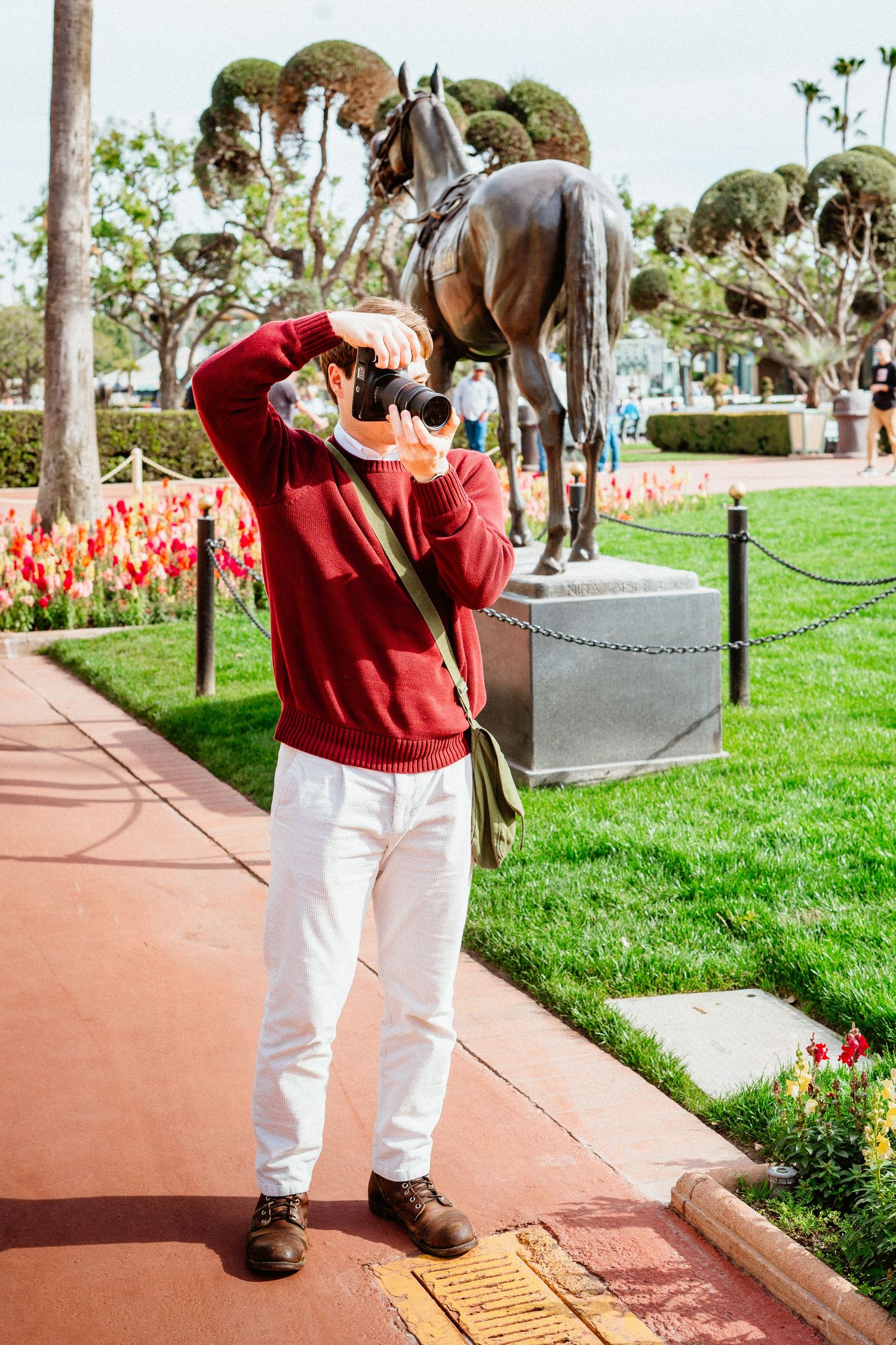 Man in red sweater and white pants using binoculars near a bronze horse statue outdoors.