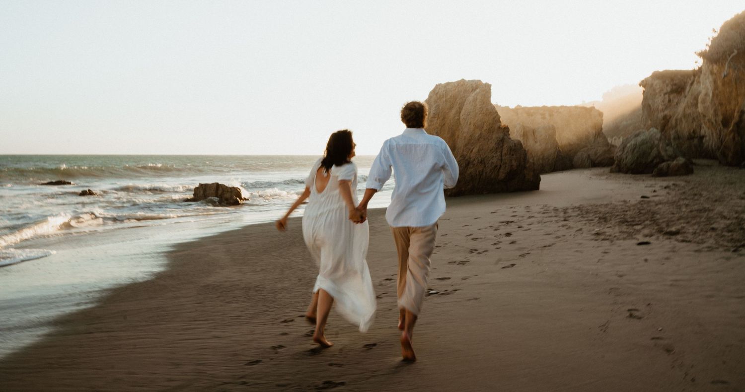 A couple in white wedding attire holds hands while running along a sandy beach at sunset near rocky cliffs.