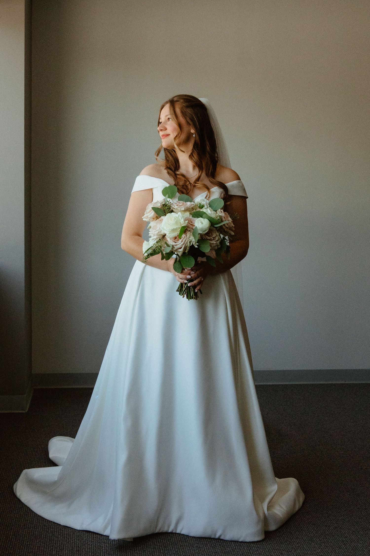 Bride in off-shoulder white A-line gown holding white rose bouquet, standing by window in elegant bridal portrait.