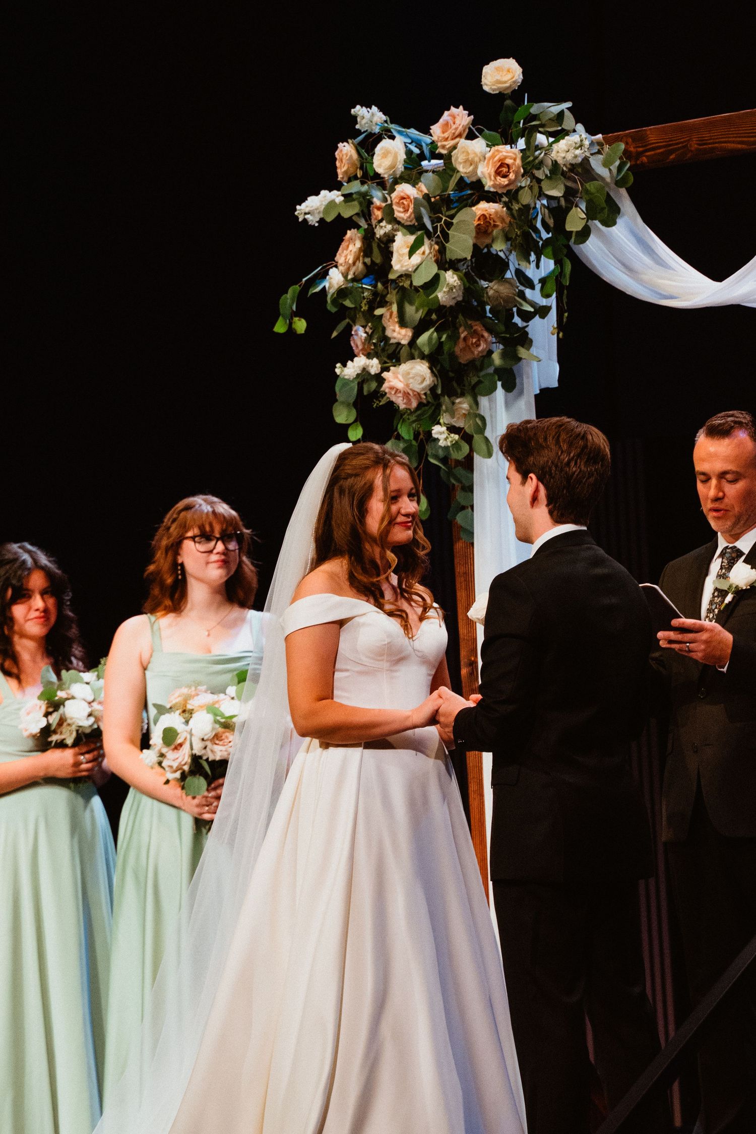 Bride smiling at groom during wedding ceremony with bridesmaids in mint dresses and floral arch backdrop.