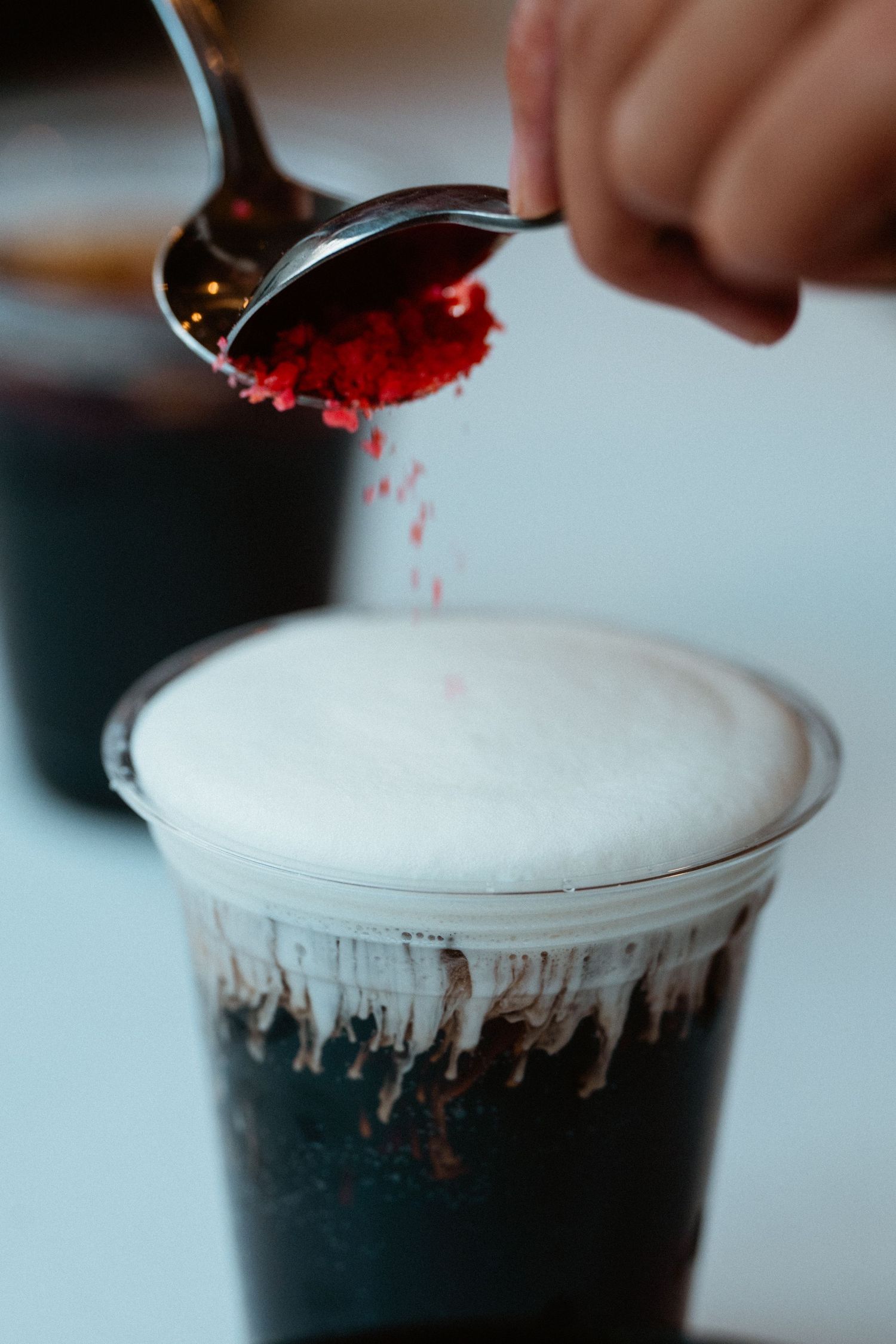 Red powder being sprinkled from a spoon onto a frothy coffee drink in a clear cup.