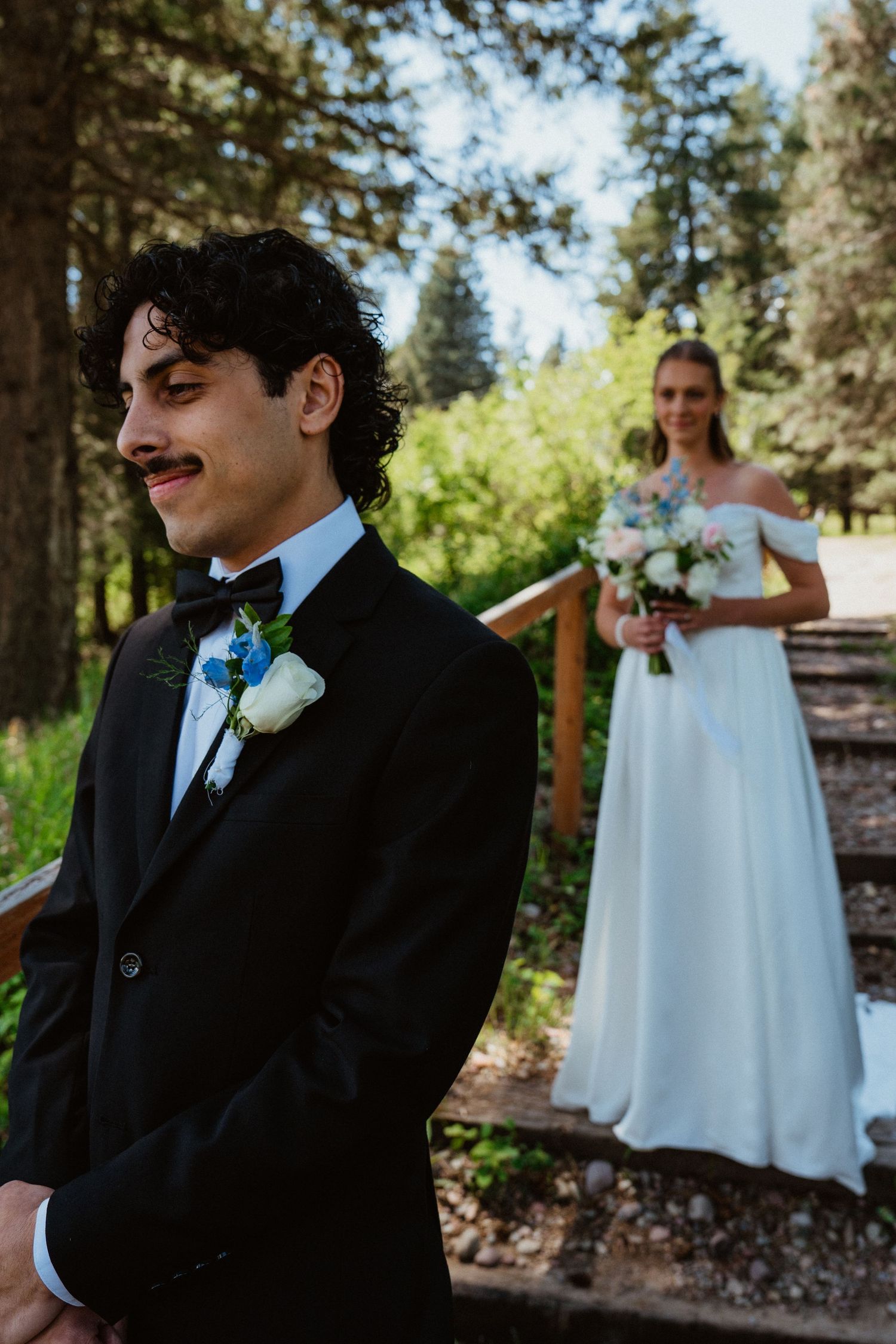 Groom in black tuxedo with boutonniere stands before bride in white off-shoulder gown holding bouquet in forest setting.