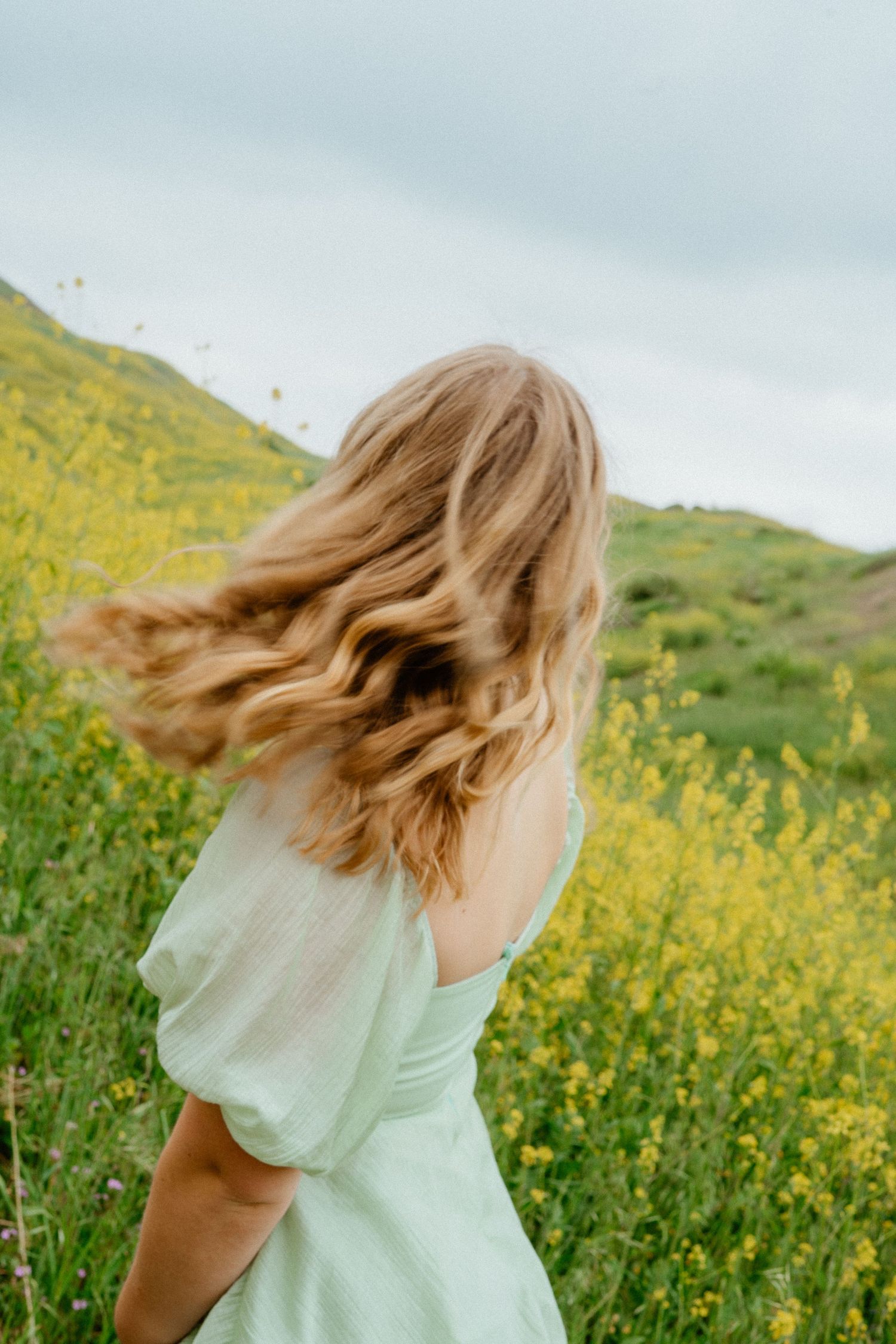 Woman in mint dress with hair blowing in wind among yellow wildflowers.