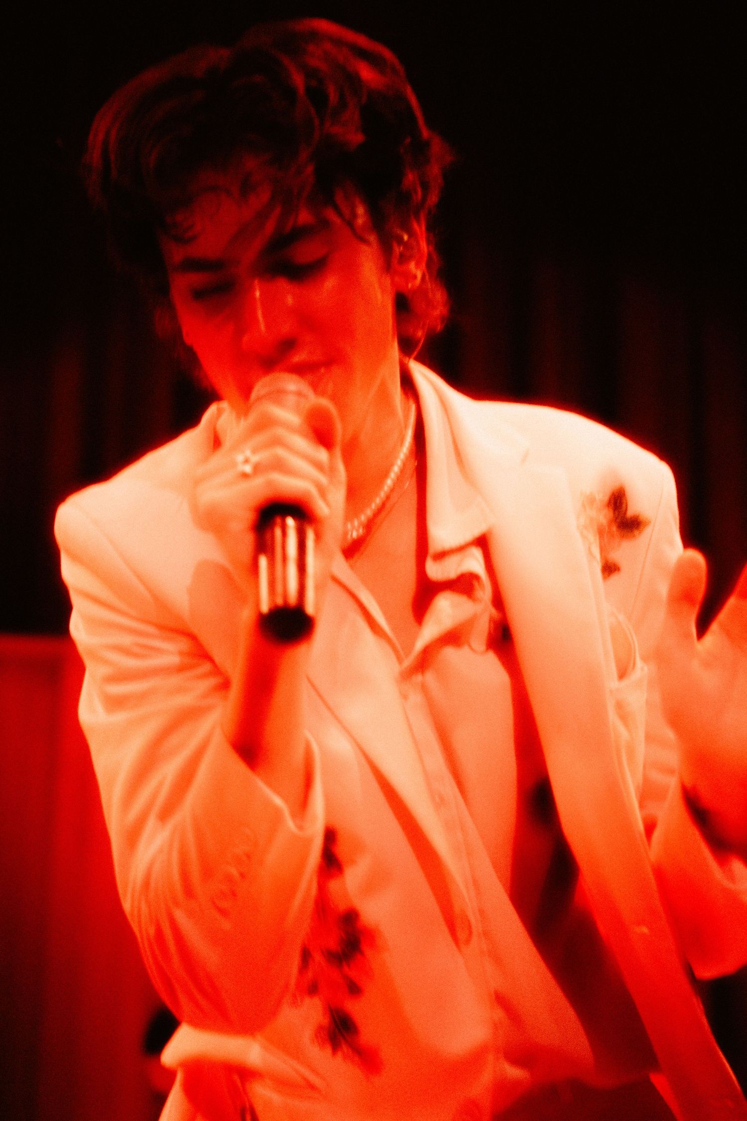 Male singer in white jacket performing with microphone under vibrant red concert stage lights.