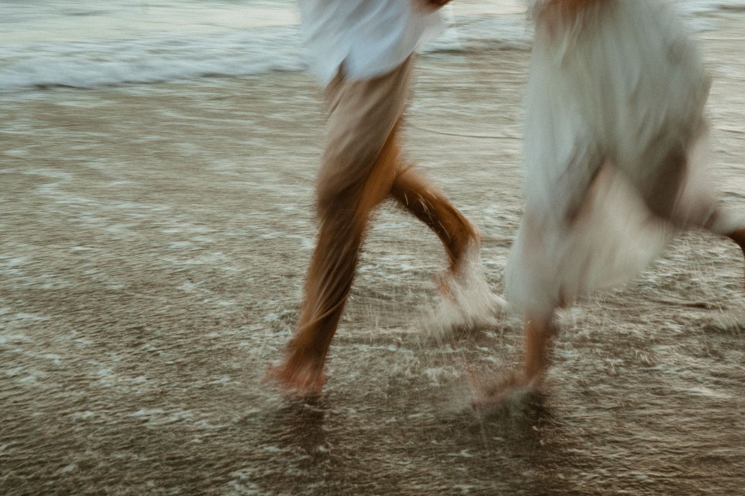 Blurred motion shot of bare feet and a dog running through shallow ocean waves on a sandy beach.