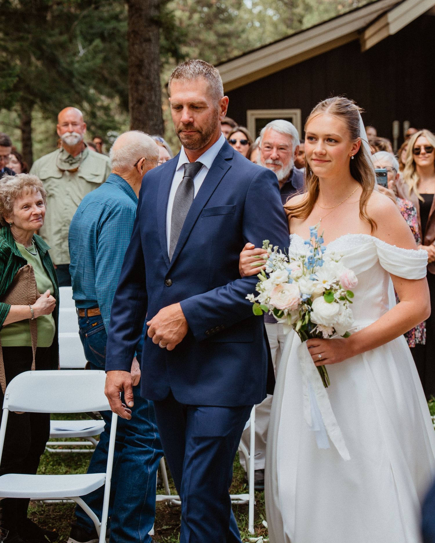 A bride in an off-shoulder white gown walks down the aisle with a man in a navy suit at an outdoor wedding.