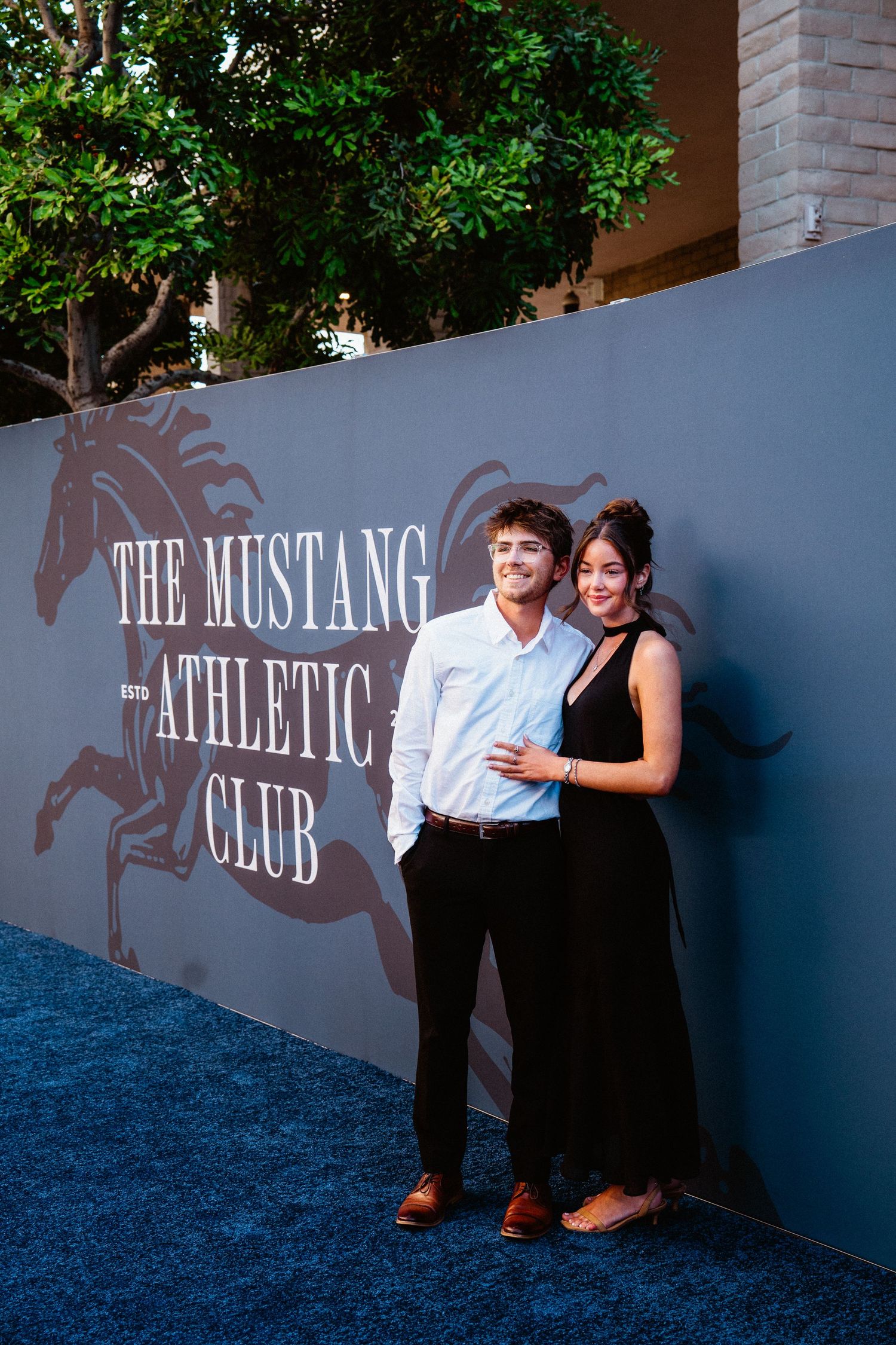 Couple posing together at The Mustang Athletic Club event on a blue carpet backdrop.