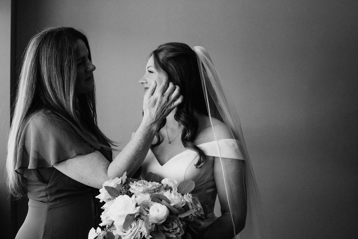 Black and white photo of a mother tenderly cupping her daughters face on her wedding day, bride holding bouquet.