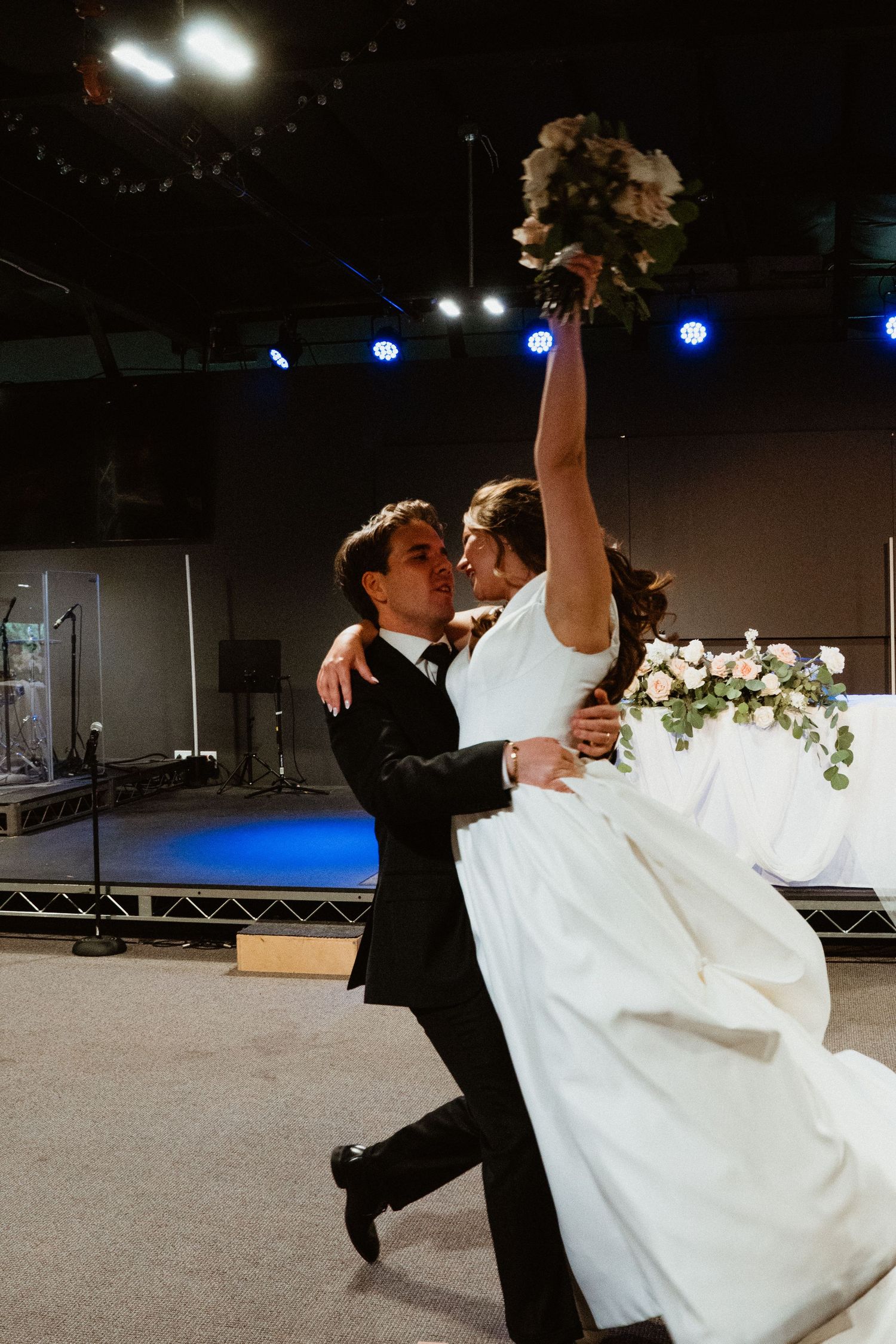 Groom lifting bride as she raises bouquet in joyful wedding celebration moment indoors.