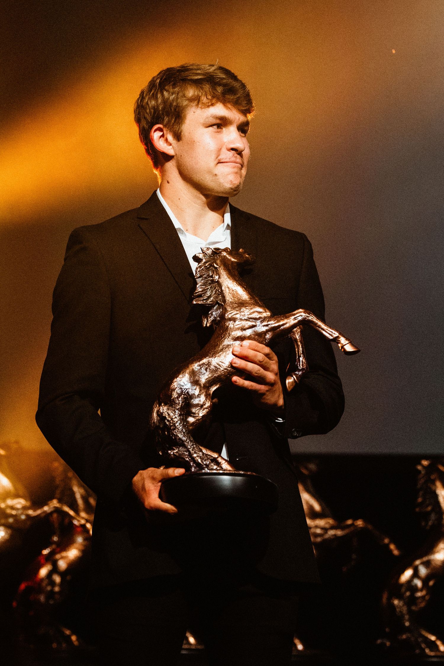 Young man in black suit holding a bronze horse trophy award on a dramatic lit stage.