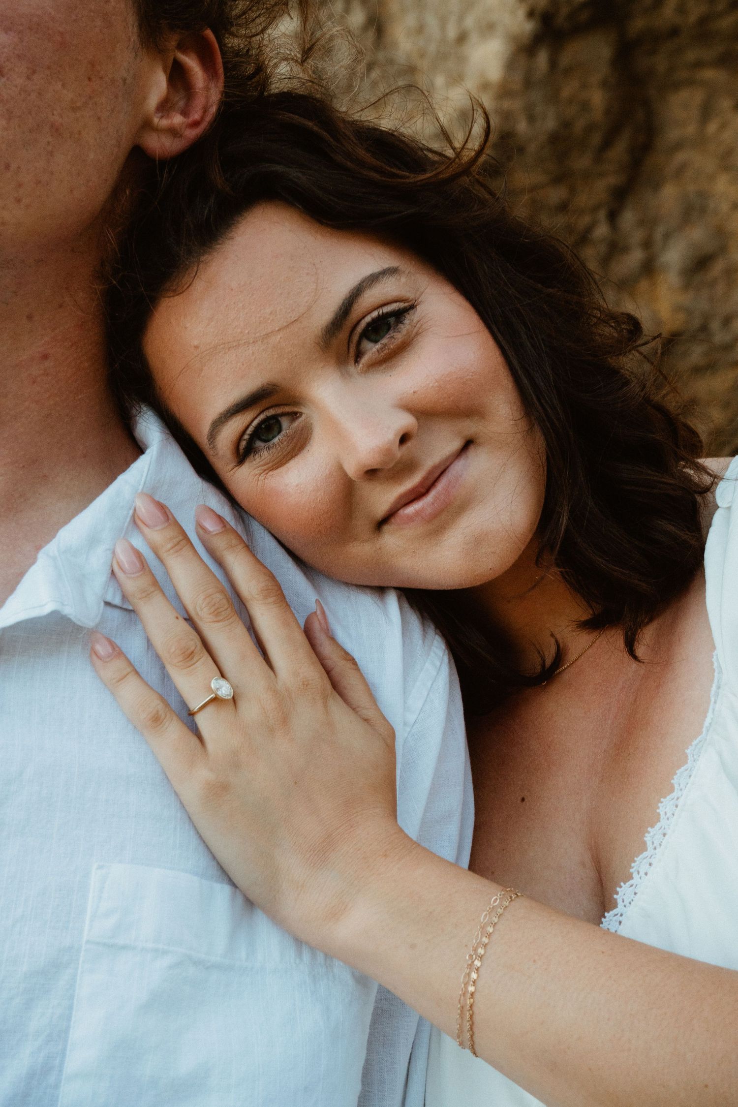 Bride leans on groom showing diamond engagement ring, smiling softly during beach engagement photo session.