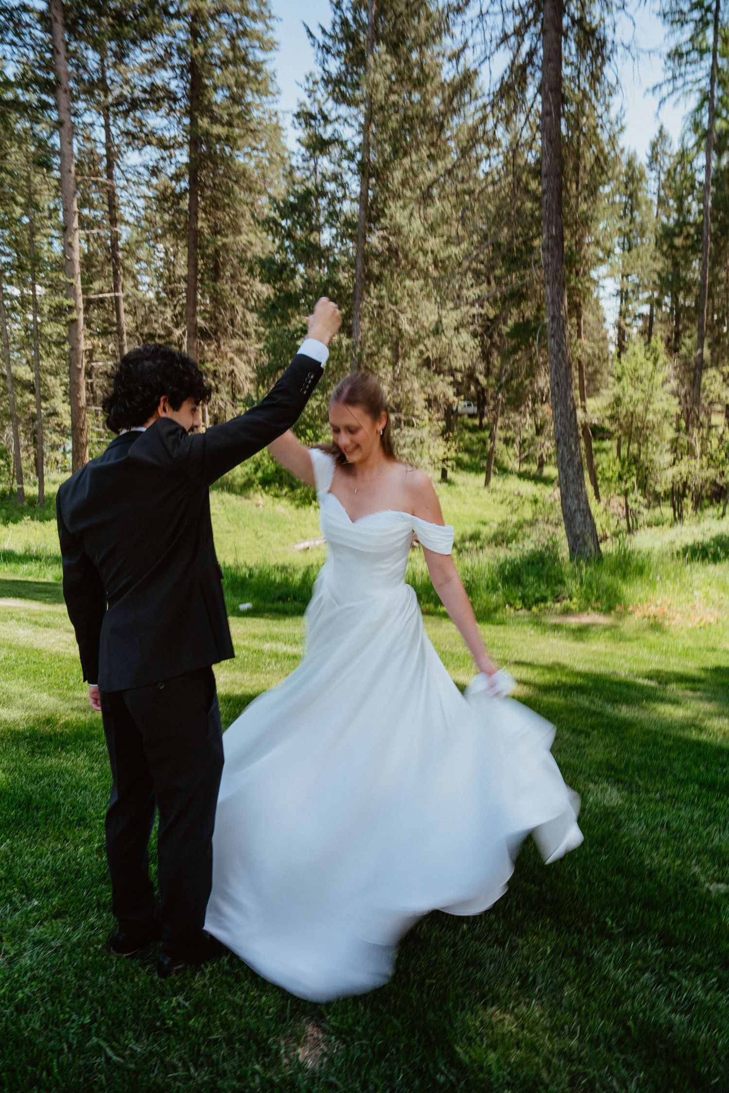 Bride in off-shoulder white gown twirls as groom spins her outdoors in a lush green forest setting.