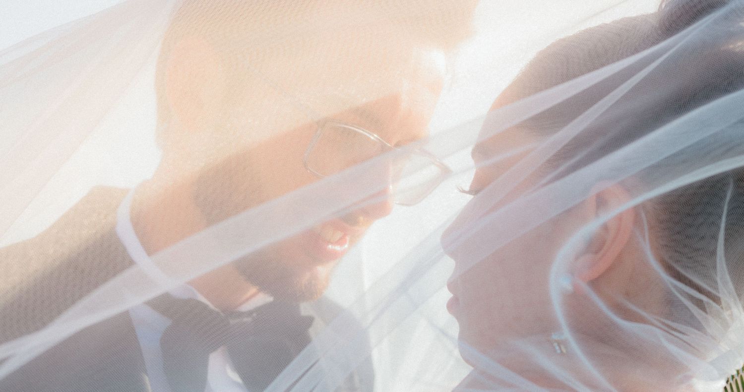 Romantic soft-focus portrait of bride and groom under sheer wedding veil in warm golden sunlight.