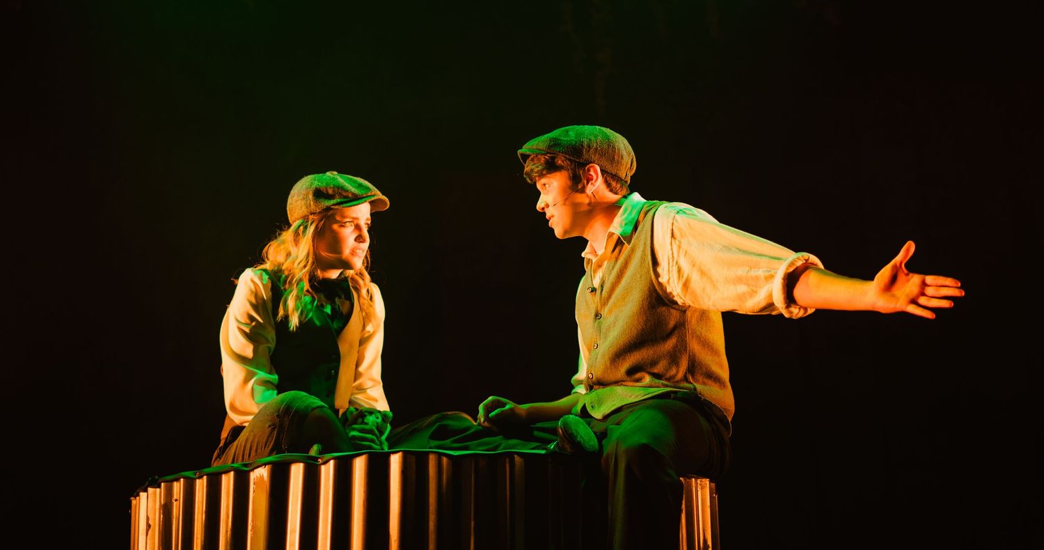 Two costumed stage performers share an intimate theatrical scene under green dramatic lighting.