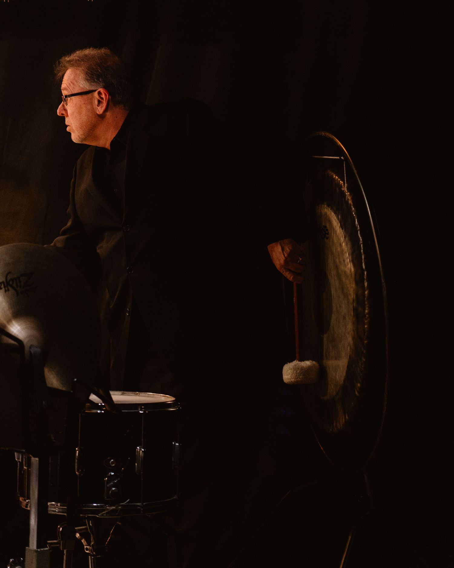 Percussionist playing a large gong with a mallet in dark concert setting.