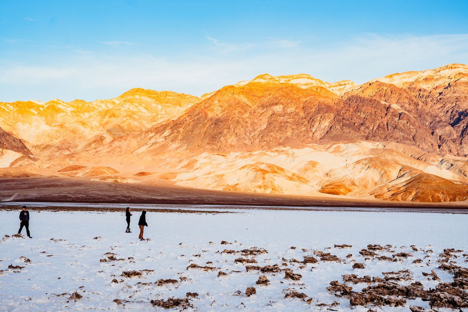 Three people walk across a vast white salt flat with dramatic golden mountain ranges at sunset.