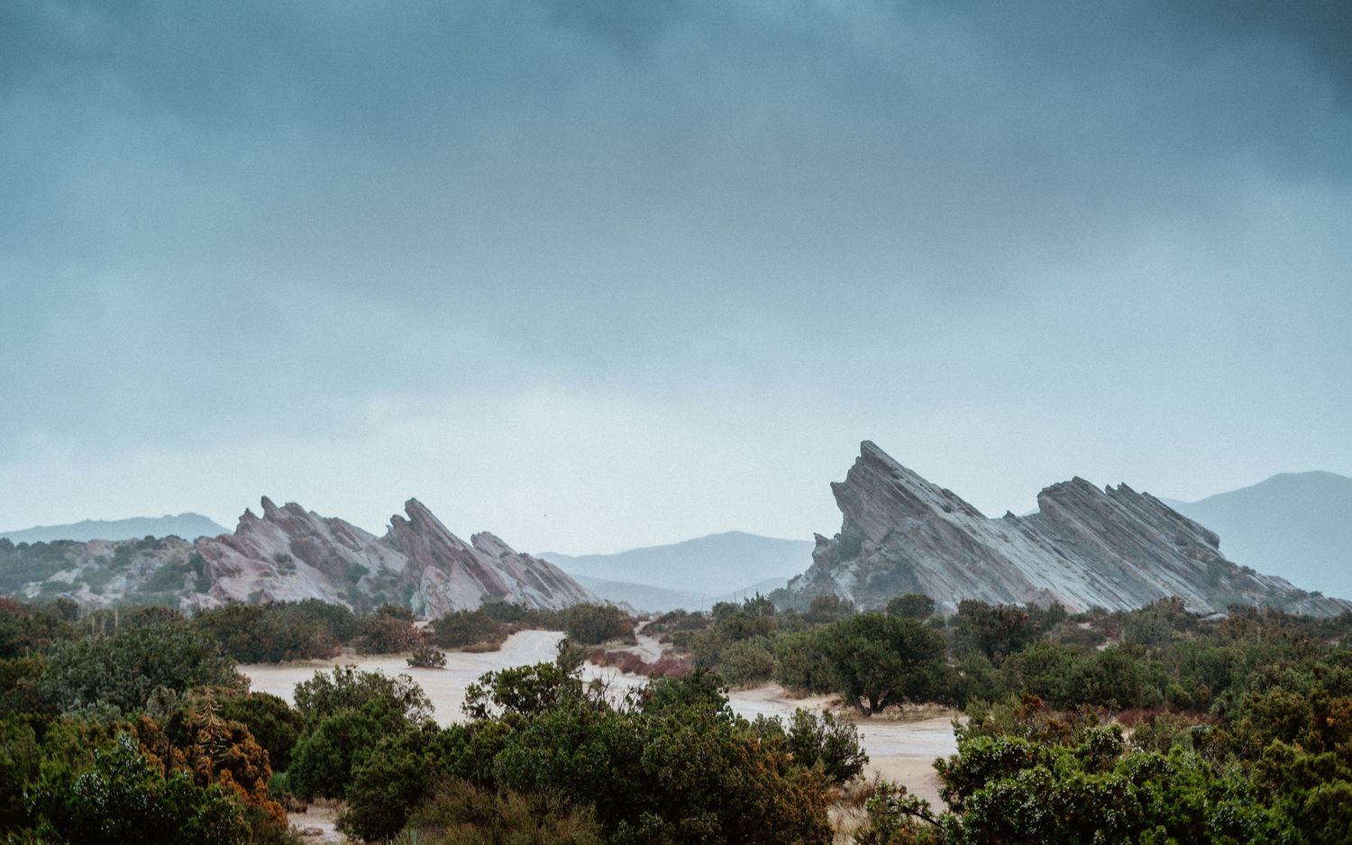Dramatic jagged rock formations at Vasquez Rocks rise above desert shrubs under moody overcast stormy sky.