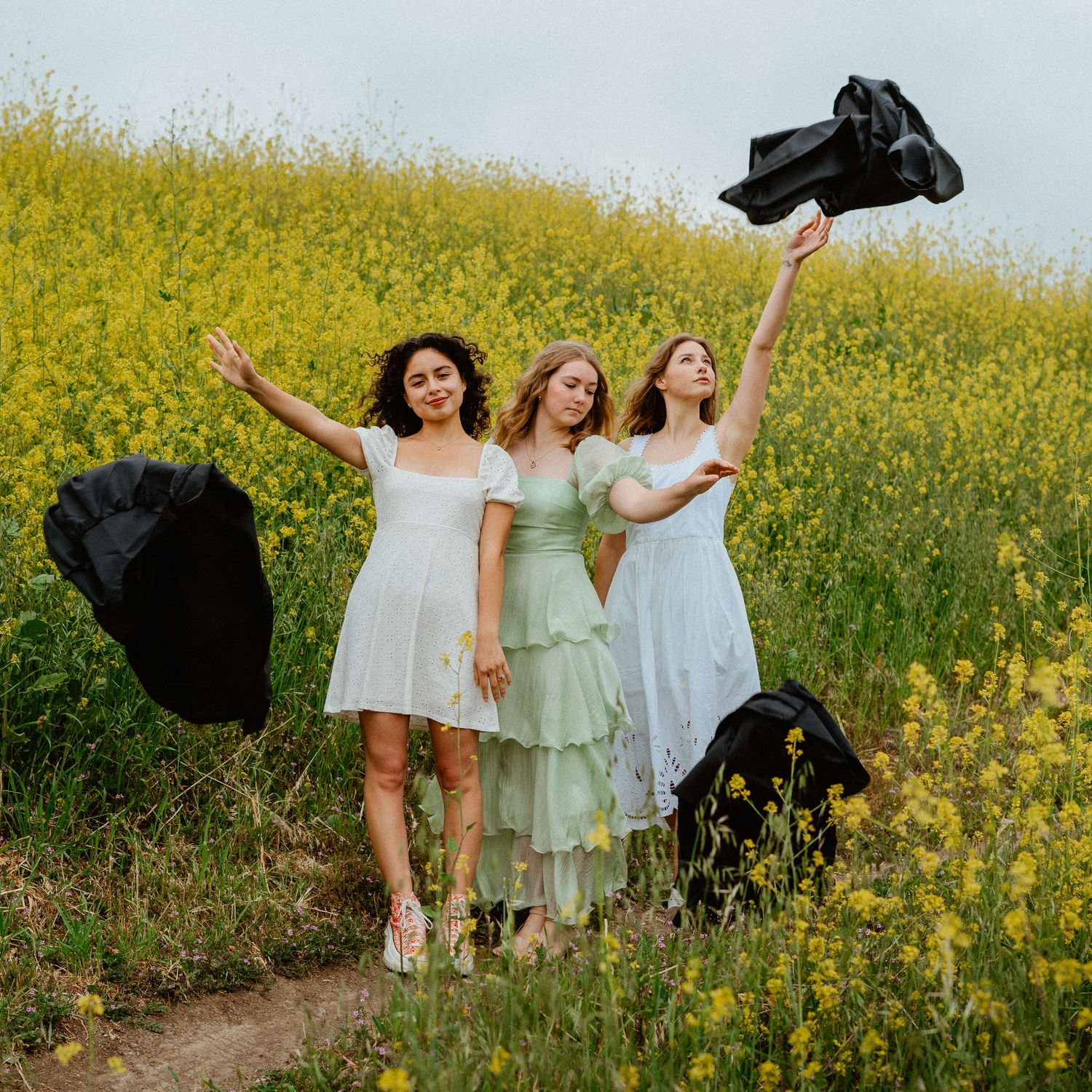 Three women in white dresses celebrating graduation by tossing black gowns in a yellow wildflower field.