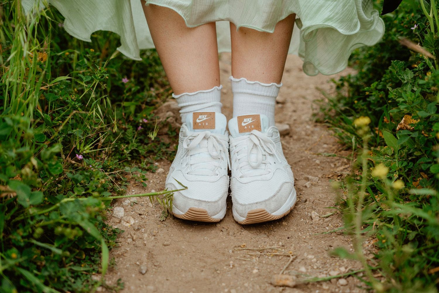 White Nike sneakers with tan soles and ankle socks worn on a dirt path surrounded by greenery.