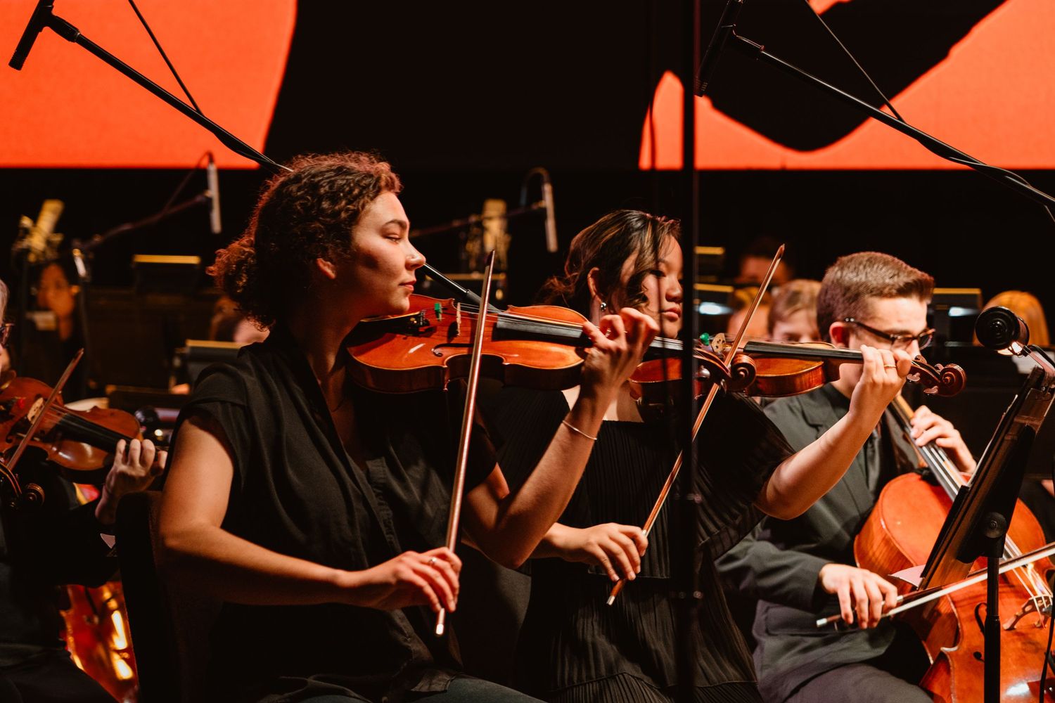 Female violinists performing in an orchestra under warm red stage lighting during a live concert.