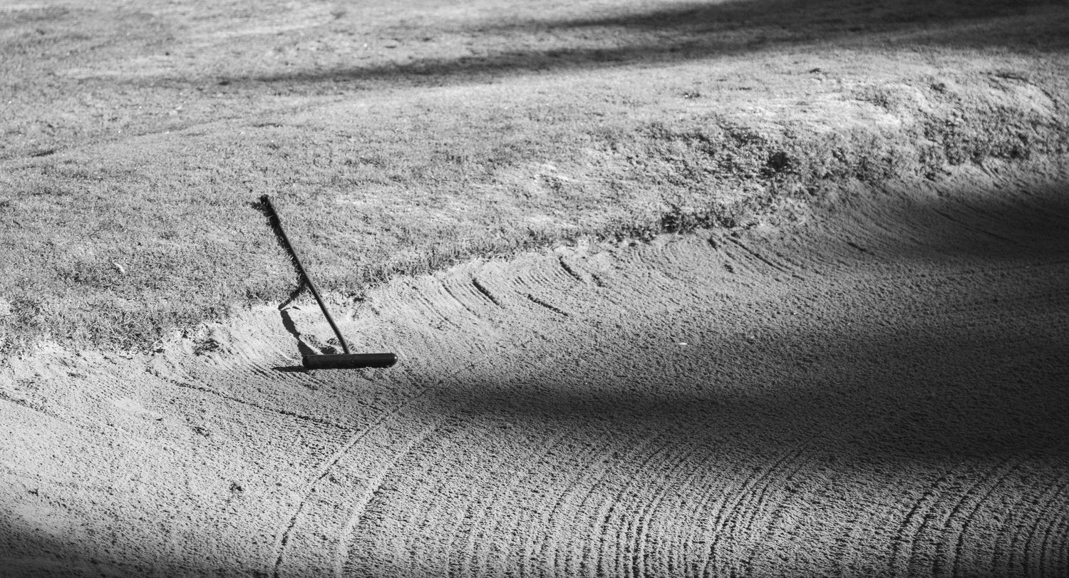 Black and white aerial view of sand bunker rake casting long shadow on textured golf course sand trap.