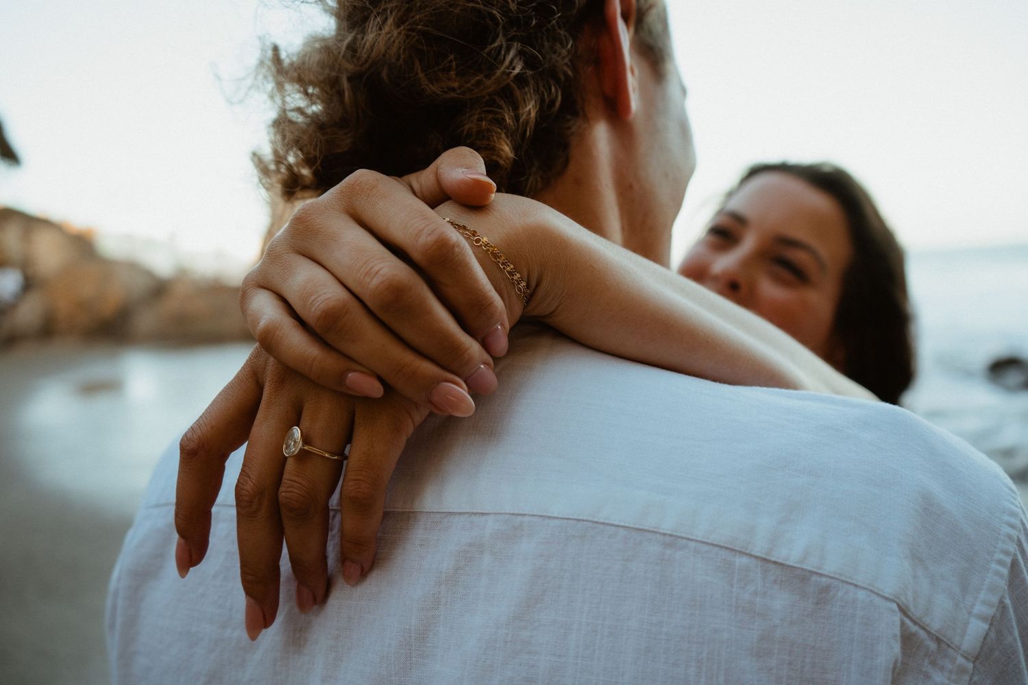 Close-up of a woman's hand with engagement ring resting on a man's shoulder at a beach.