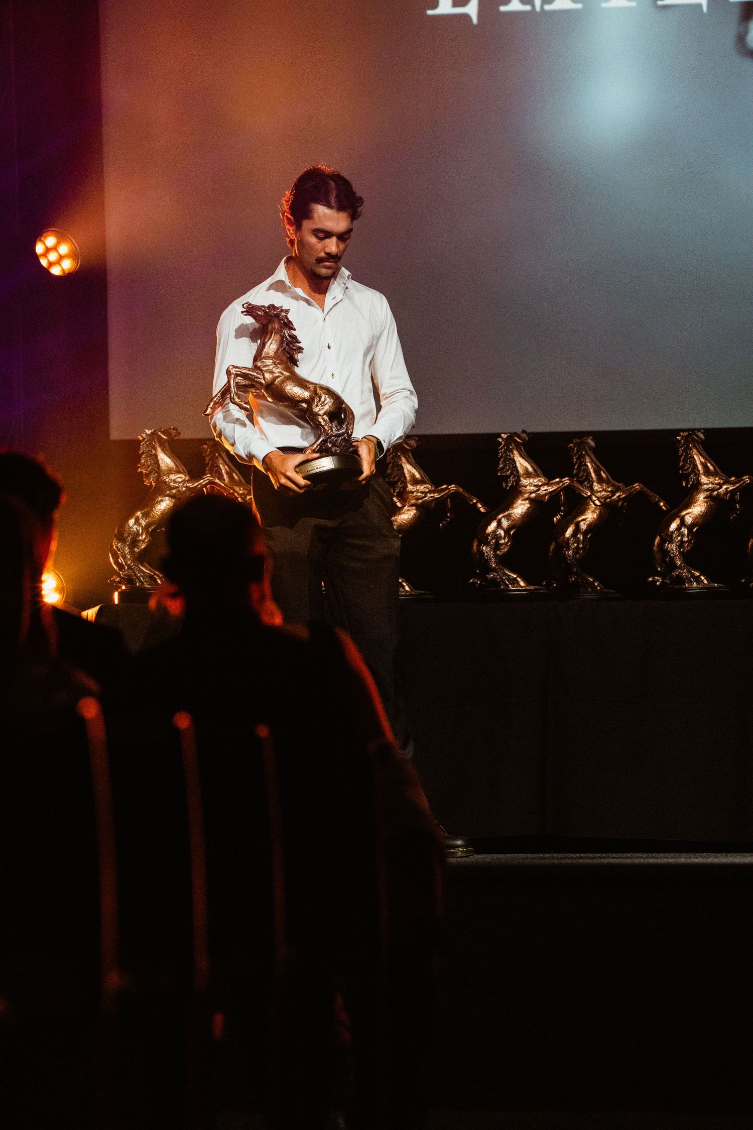 Man in white shirt holding a golden horse trophy on stage at an awards ceremony with dramatic lighting.