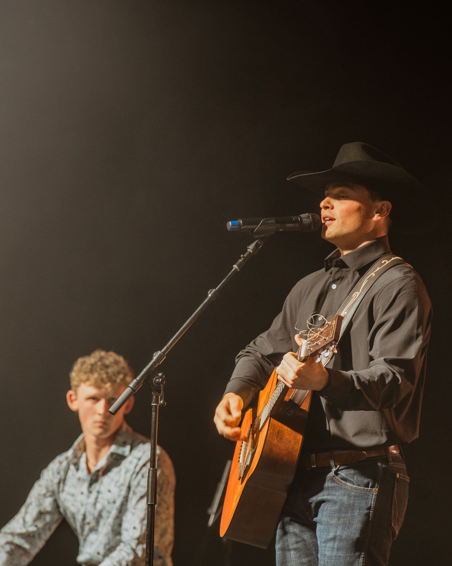 A country singer in a black hat plays acoustic guitar and sings at a live performance on stage.