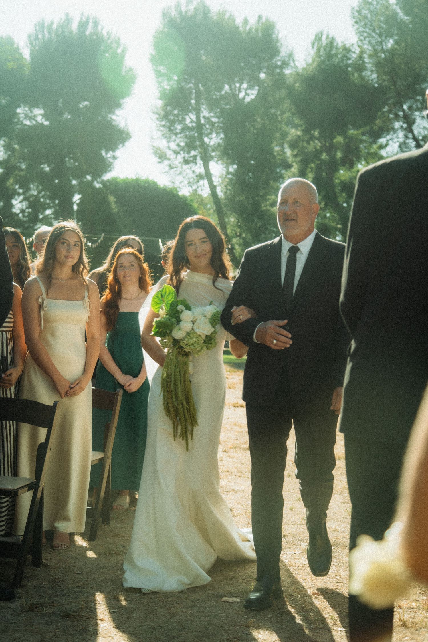 Bride walking down outdoor aisle with father, bridesmaids in green dresses, surrounded by tall trees in sunlit ceremony.