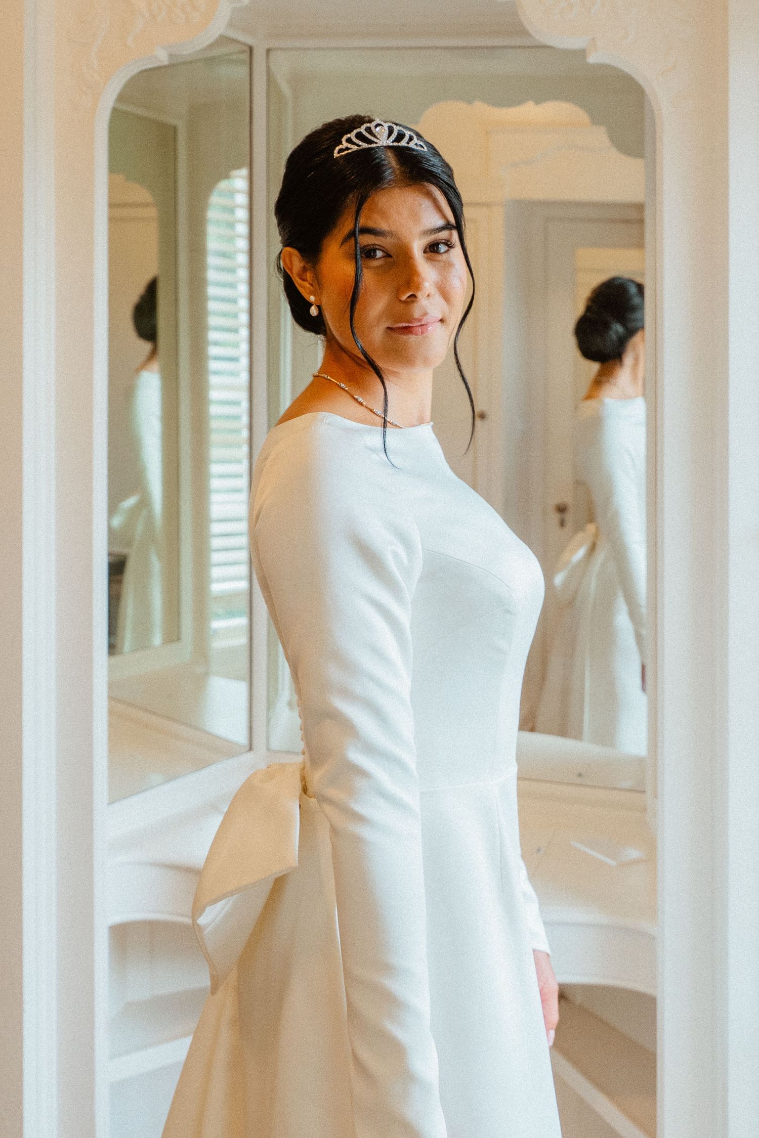 Elegant bride in long-sleeve white gown with tiara poses in front of ornate mirror before wedding ceremony.