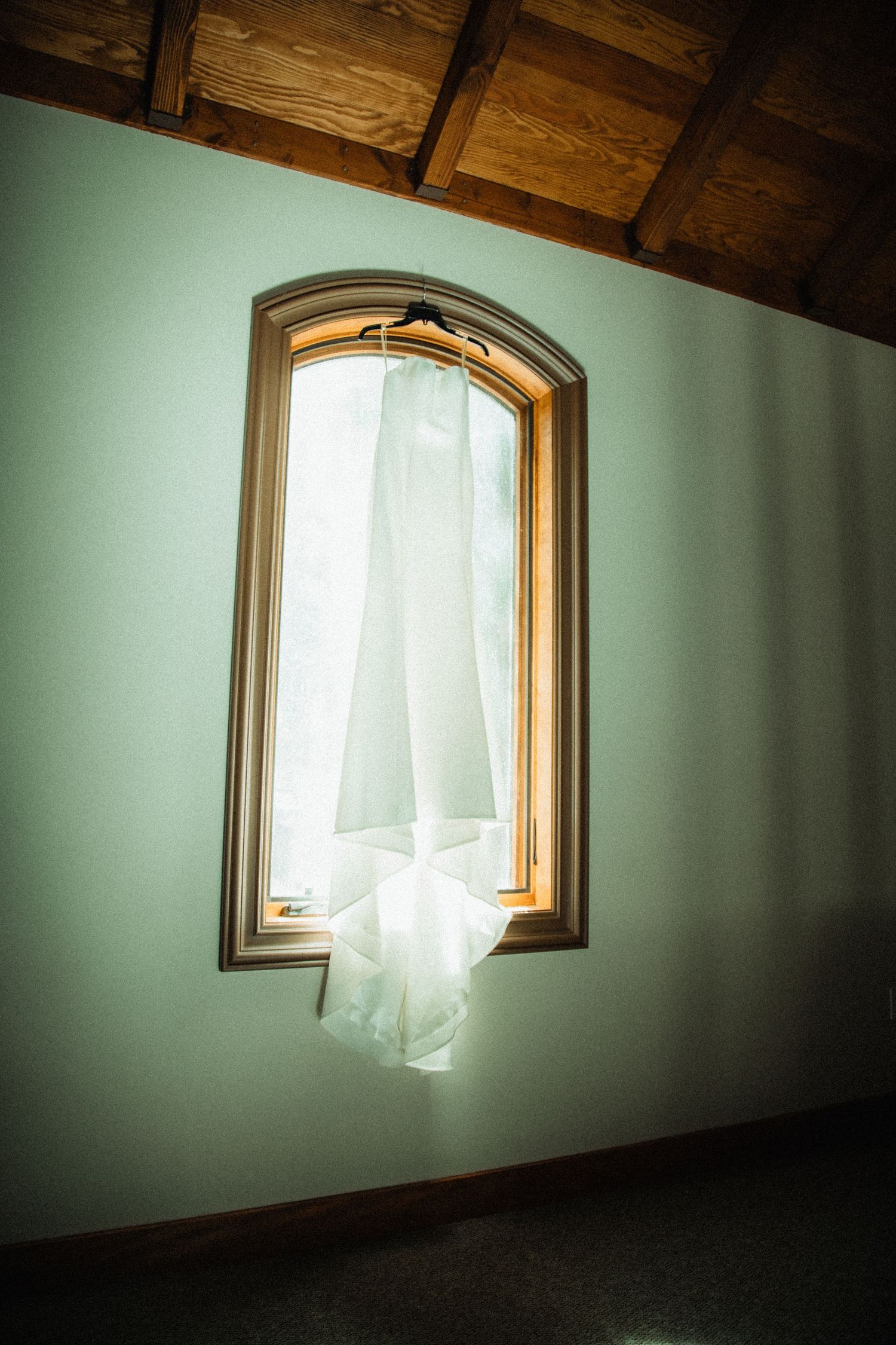 Elegant white wedding dress hanging in arched window with natural light in rustic wood-ceiling venue.