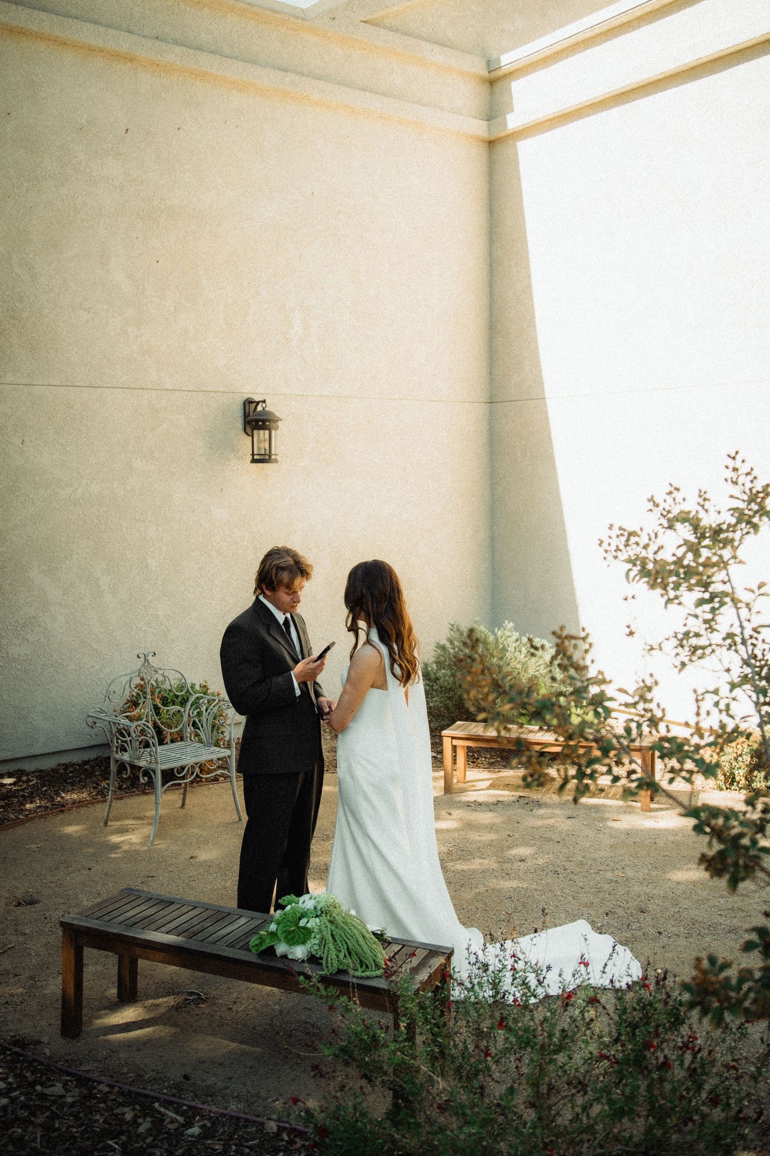 Bride and groom exchanging vows outdoors at sunset against white stucco wall at wedding ceremony.
